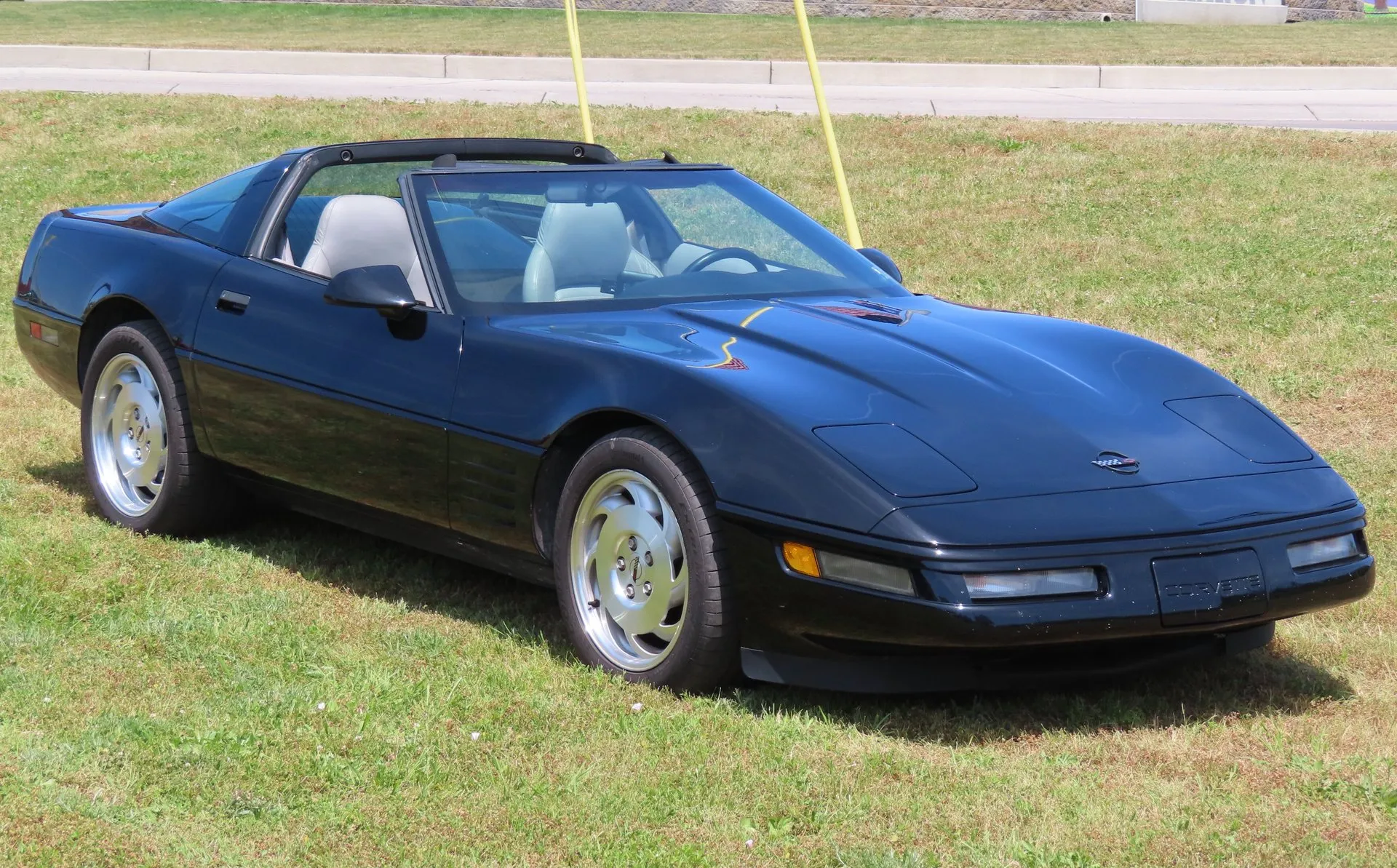 Black C4 Corvette coupe with its roof panel off, parked on grass and seen from the front right, with a light gray leather interior, silver slotted wheels and straight horizontal strakes behind the front wheel.