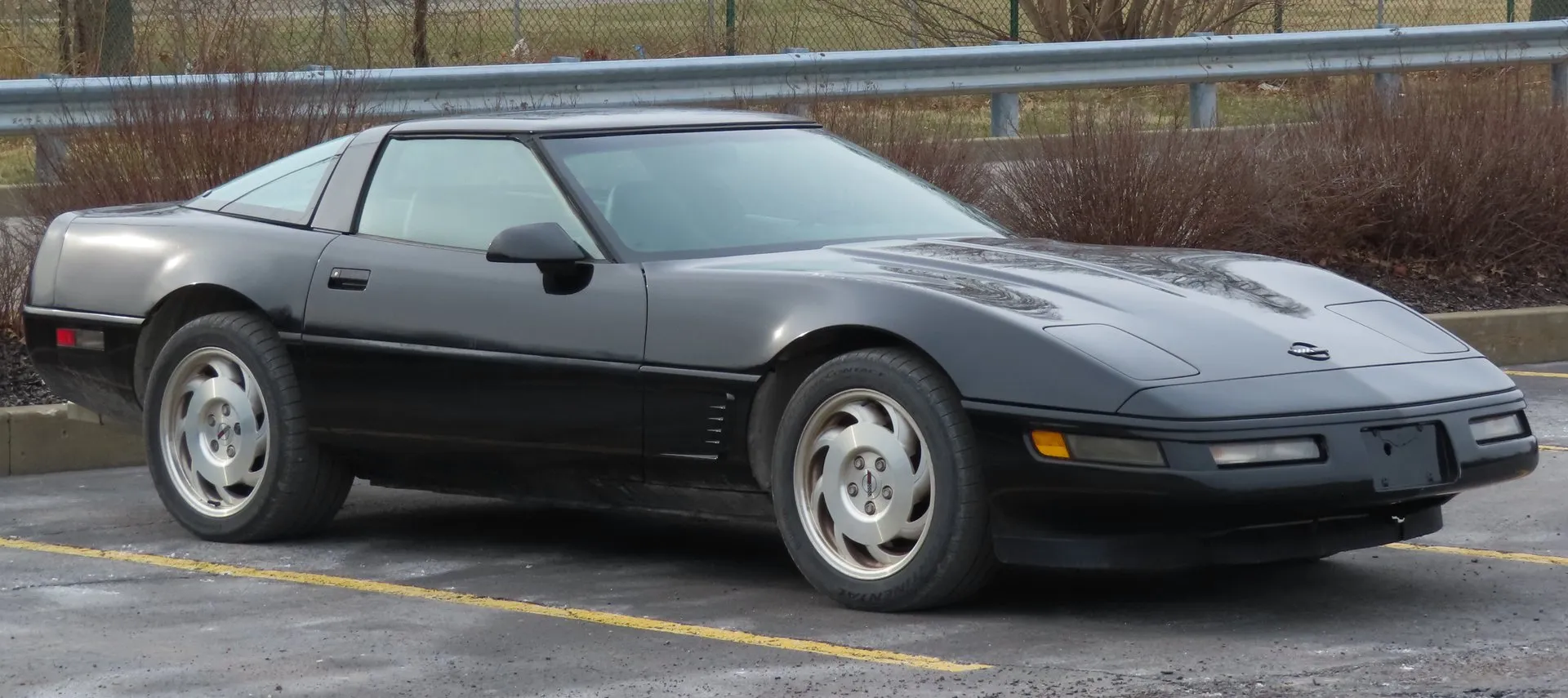 Black C4 Corvette coupe in a parking lot, seen from the front right, with a black roof, silver slotted wheels and a vented gill panel behind the front wheel.