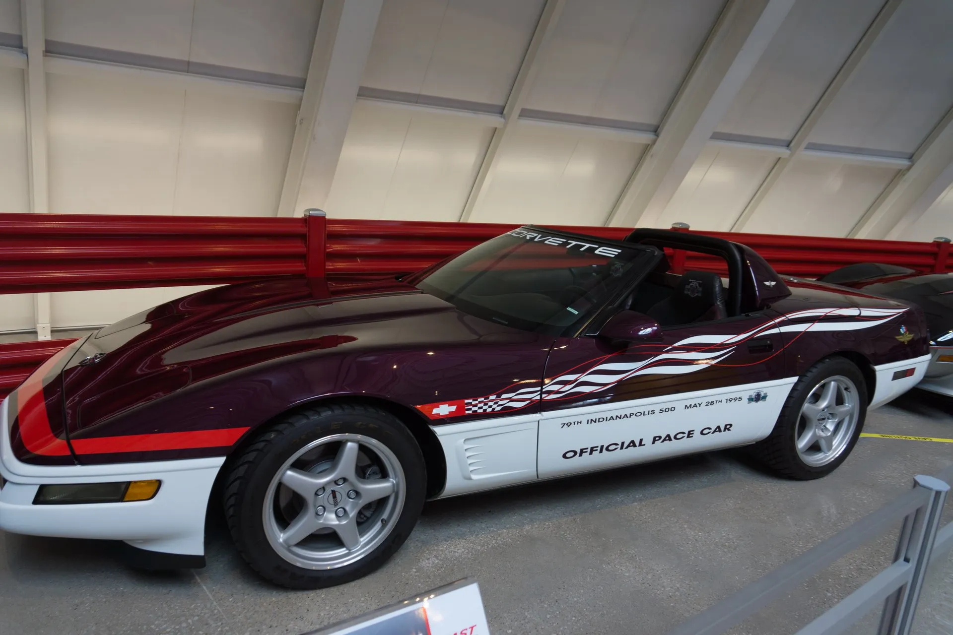 Dark purple and white C4 Corvette convertible with its top down and a black roll hoop behind the seats, in a museum hall, seen from the left side, with door lettering reading 79th Indianapolis 500 May 28th 1995 Official Pace Car and silver five-spoke wheels.