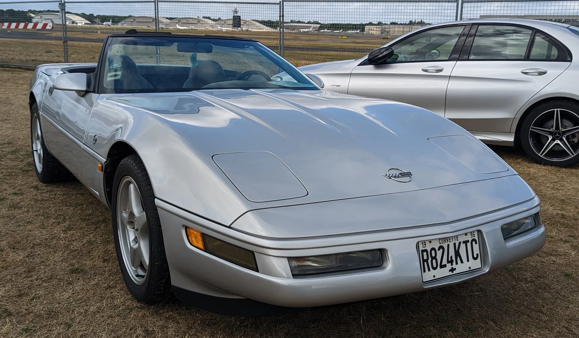 Silver C4 Corvette convertible with its top down and silver five-spoke wheels, parked on dry grass at a show beside a silver sedan, seen from the front right.