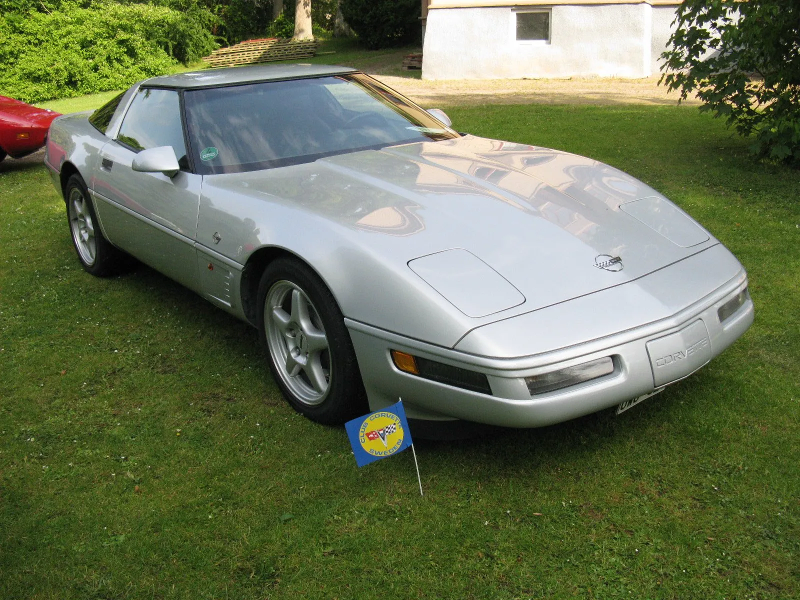 Silver C4 Corvette coupe on silver five-spoke wheels, parked on grass with a small Club Corvette Sweden flag at its front wheel, seen from the front right.