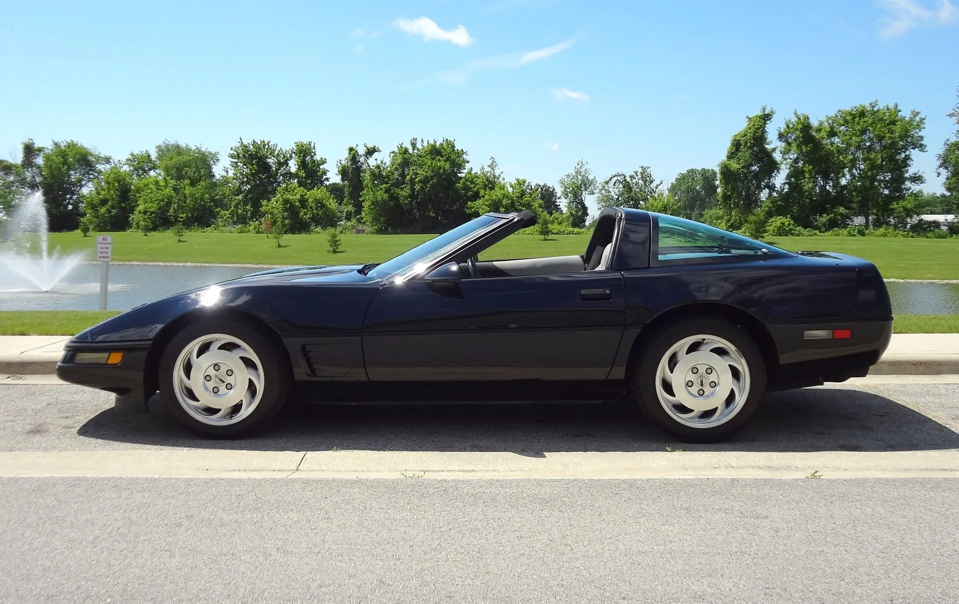 Black C4 Corvette coupe with its roof panel removed, parked at a curb beside a pond with a fountain, seen square from the left side, showing the angled vents behind the front wheel.