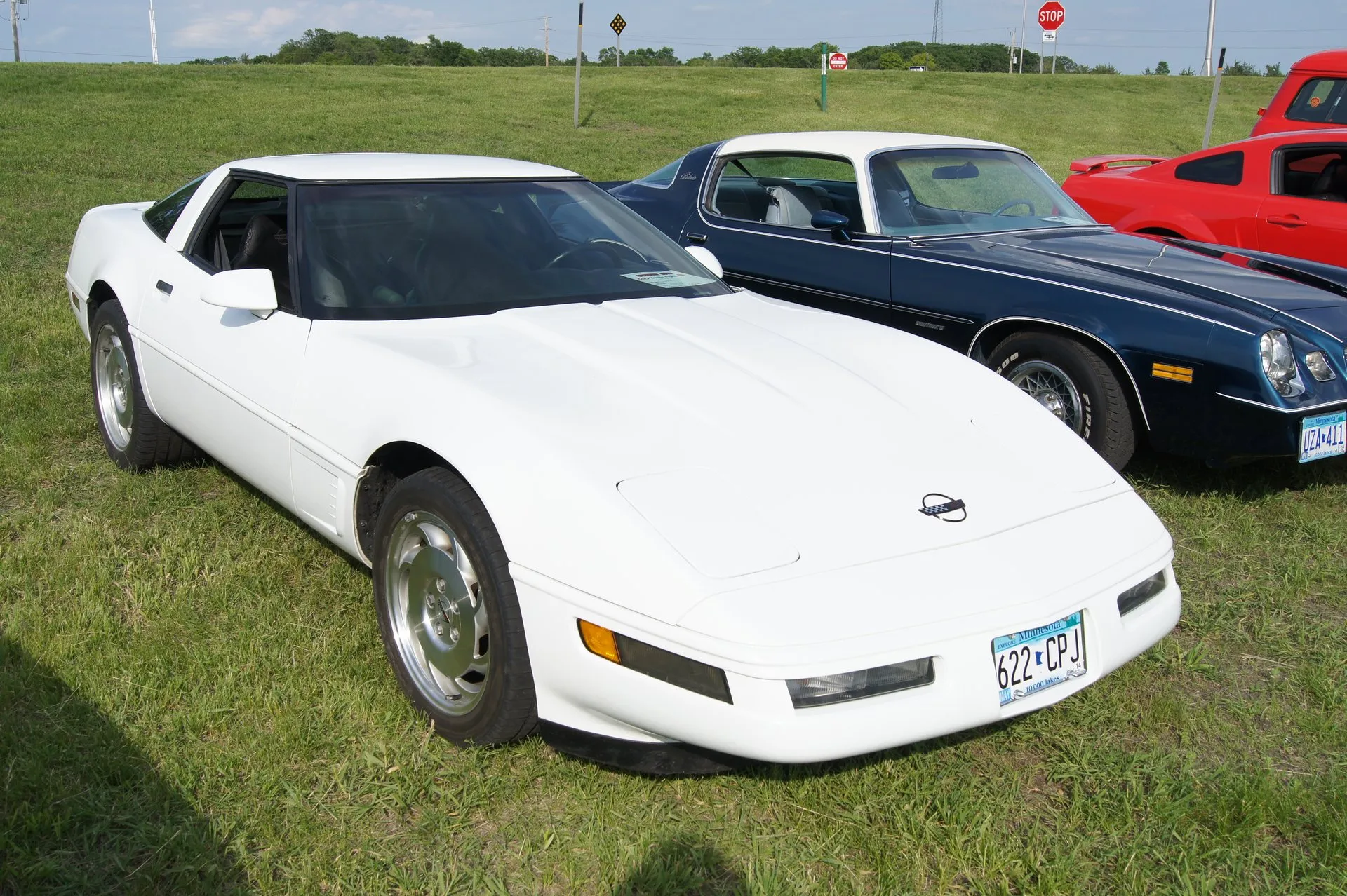 White C4 Corvette coupe on silver wheels, parked on grass at a car show, seen from the front right, with a dark blue Camaro beside it.