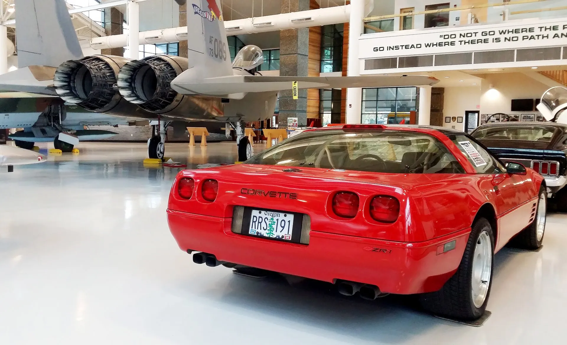 Red C4 Corvette ZR-1 coupe seen from behind and to its right in an aviation museum hall, with a ZR-1 emblem on the right of the rear fascia, raised CORVETTE letters, square taillamps, a stop lamp at the top of the rear glass and a jet fighter behind it.