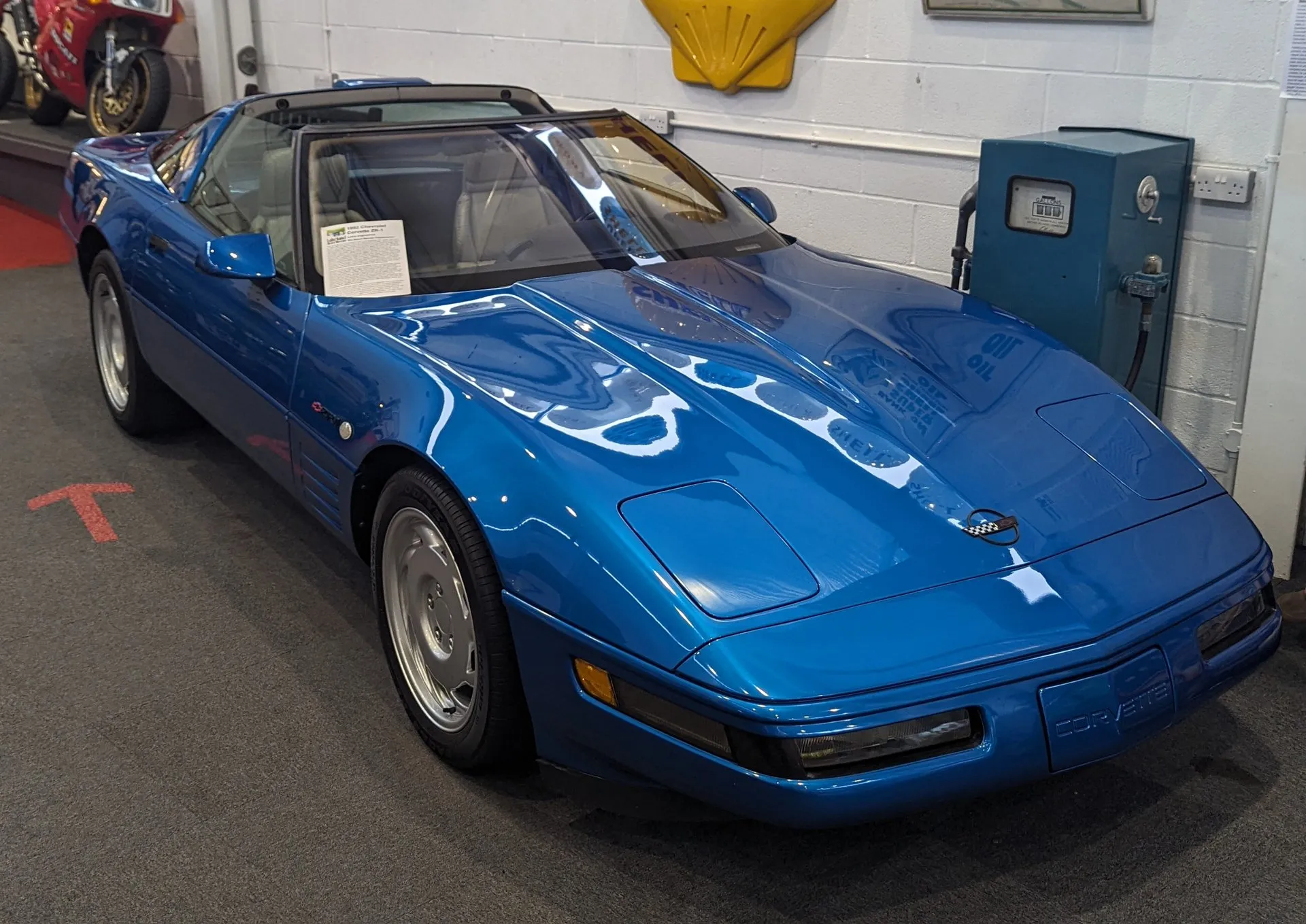 Blue C4 Corvette ZR-1 coupe with its roof panel off, seen from the front left in a museum, with a ZR-1 emblem above the vents behind the front wheel, a placard on the dashboard and an old garage pump against the wall behind.