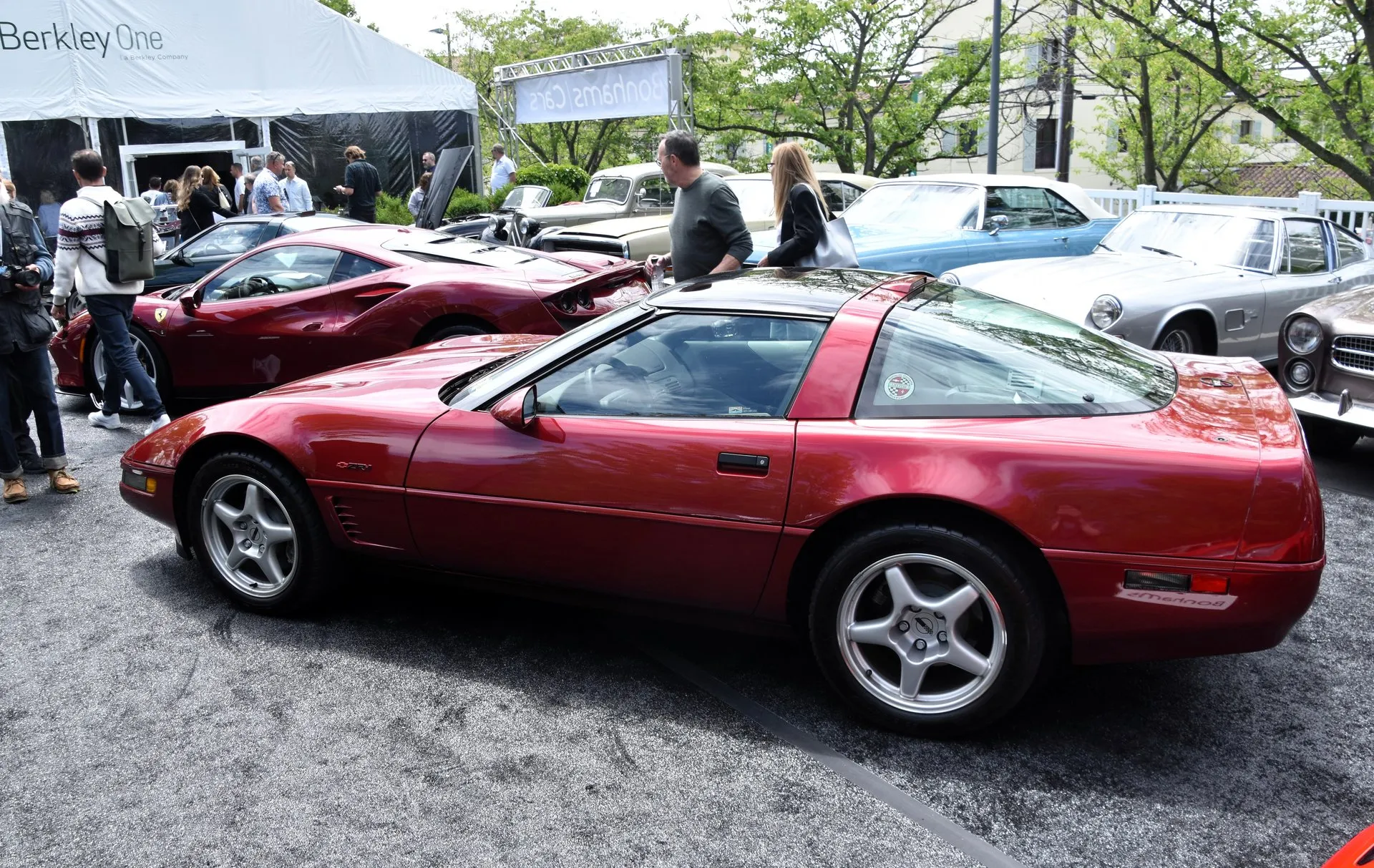 Dark red C4 Corvette ZR-1 coupe seen from its left side at an outdoor classic car sale, with a ZR-1 emblem above the vents behind the front wheel and silver five-spoke wheels, a red Ferrari and other classic cars behind it.