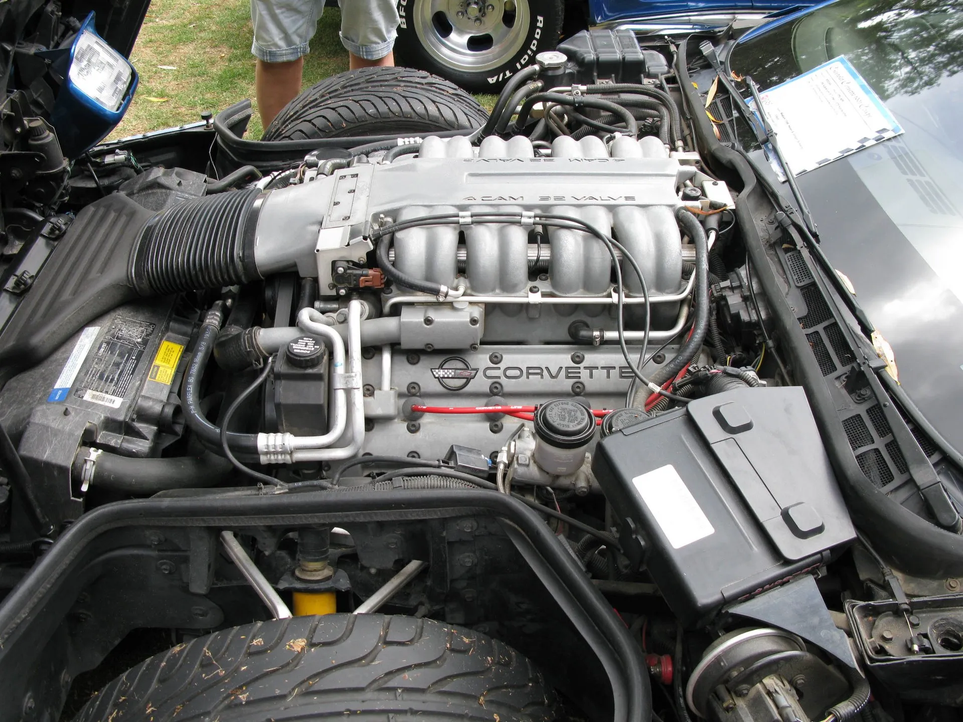 Engine bay of a blue C4 Corvette with the clamshell hood raised, showing a silver intake plenum lettered 4 CAM 32 VALVE above its intake runners, a cam cover lettered CORVETTE, and a front tire at the bottom of the frame.