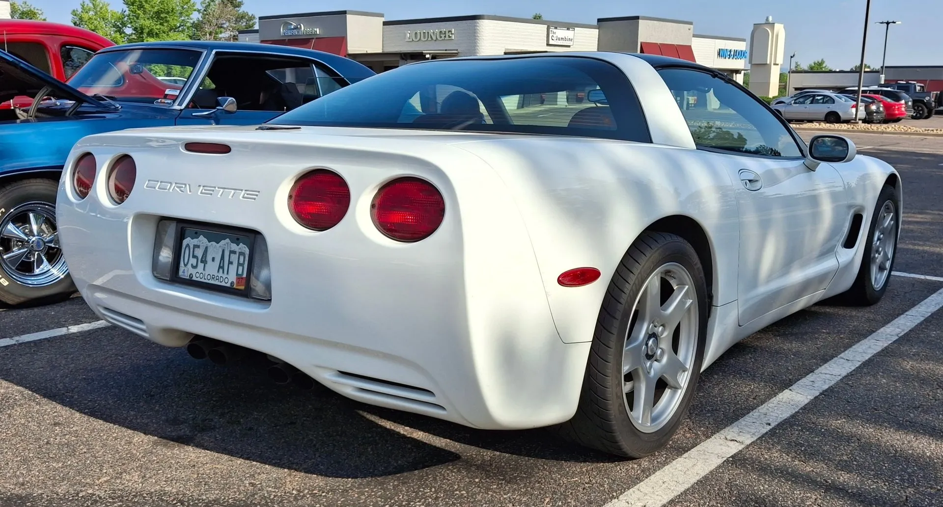 White C5 Corvette coupe parked in a car show lot, seen from the right rear, with four round red taillights, Corvette lettering across the rear, silver five-spoke wheels and a Colorado license plate, a blue classic car beside it and shops behind.