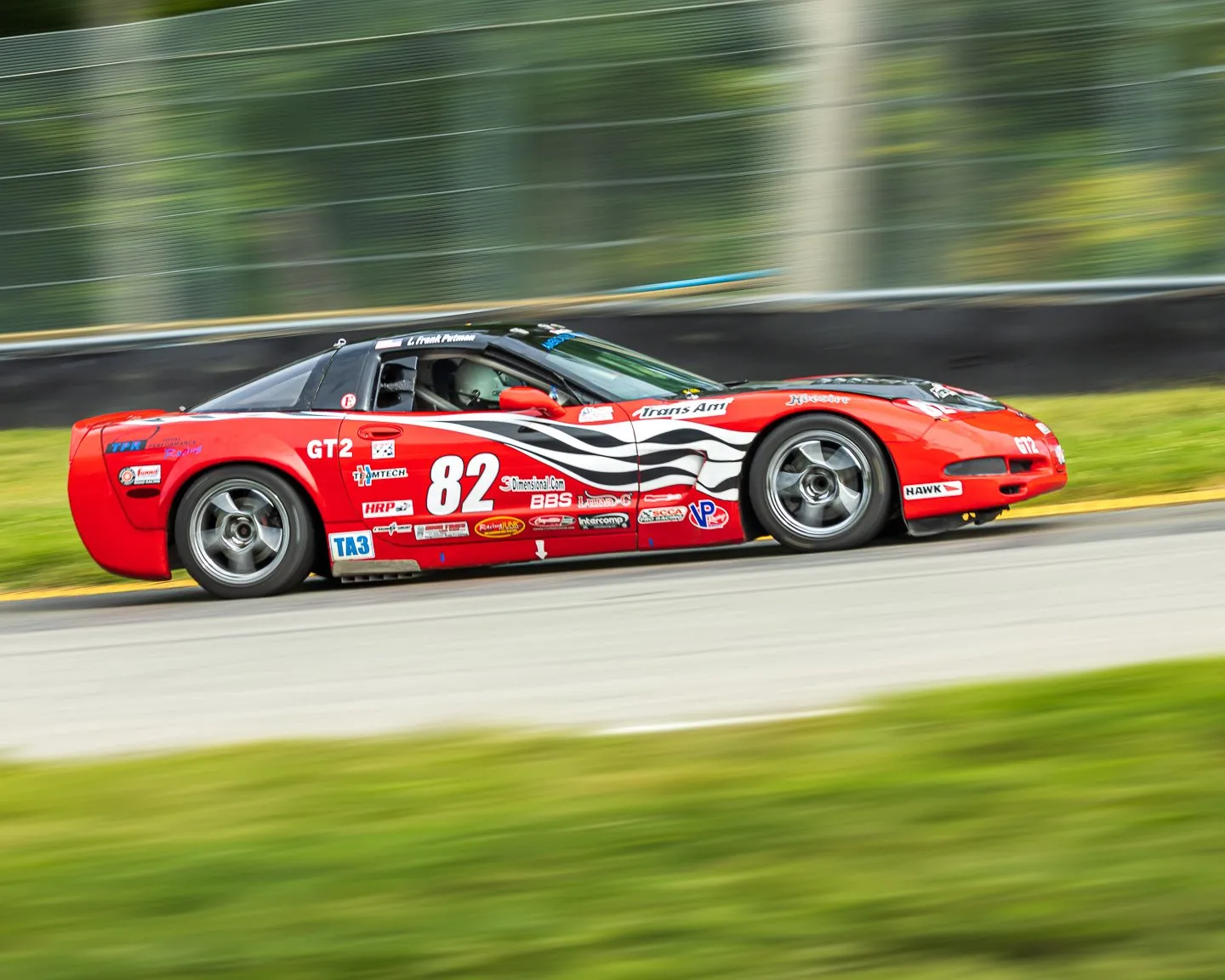 Red C5 Corvette coupe with white and black side graphics, sponsor decals and the number 82 racing past a catch fence, shot as a pan so the background blurs, with a helmeted driver inside.