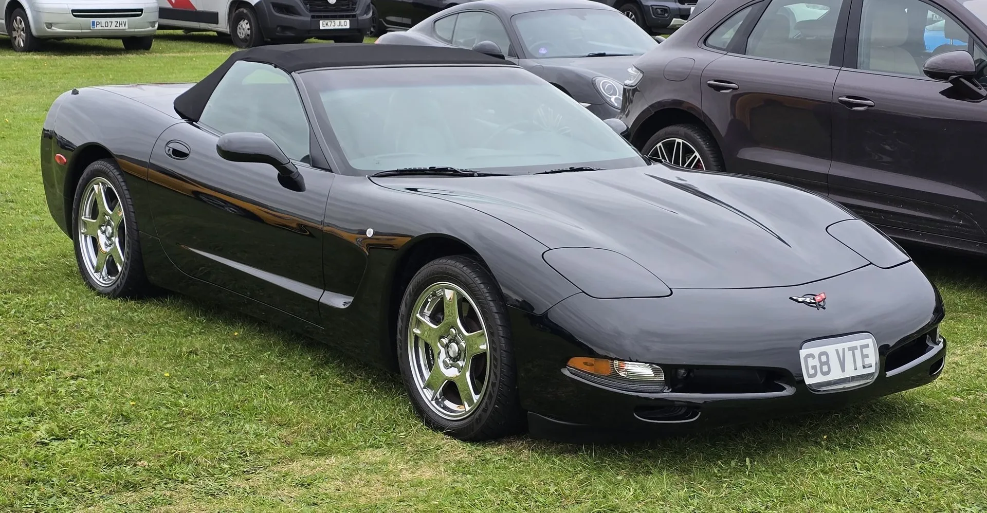 Black C5 Corvette convertible with its black cloth top up, parked on grass at a car show and seen from the front left, with chrome-finish five-spoke wheels and a British license plate.
