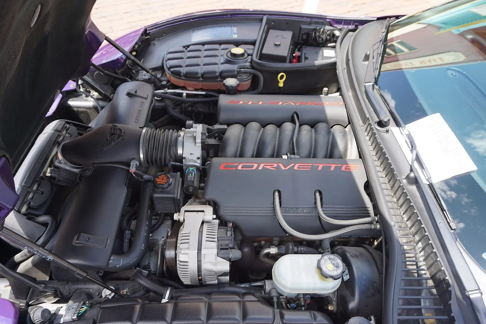 Engine bay of a 1998 C5 Corvette with the hood up; at the far corner by the windshield, the battery with a red positive terminal cover sits beside the black underhood fuse box.