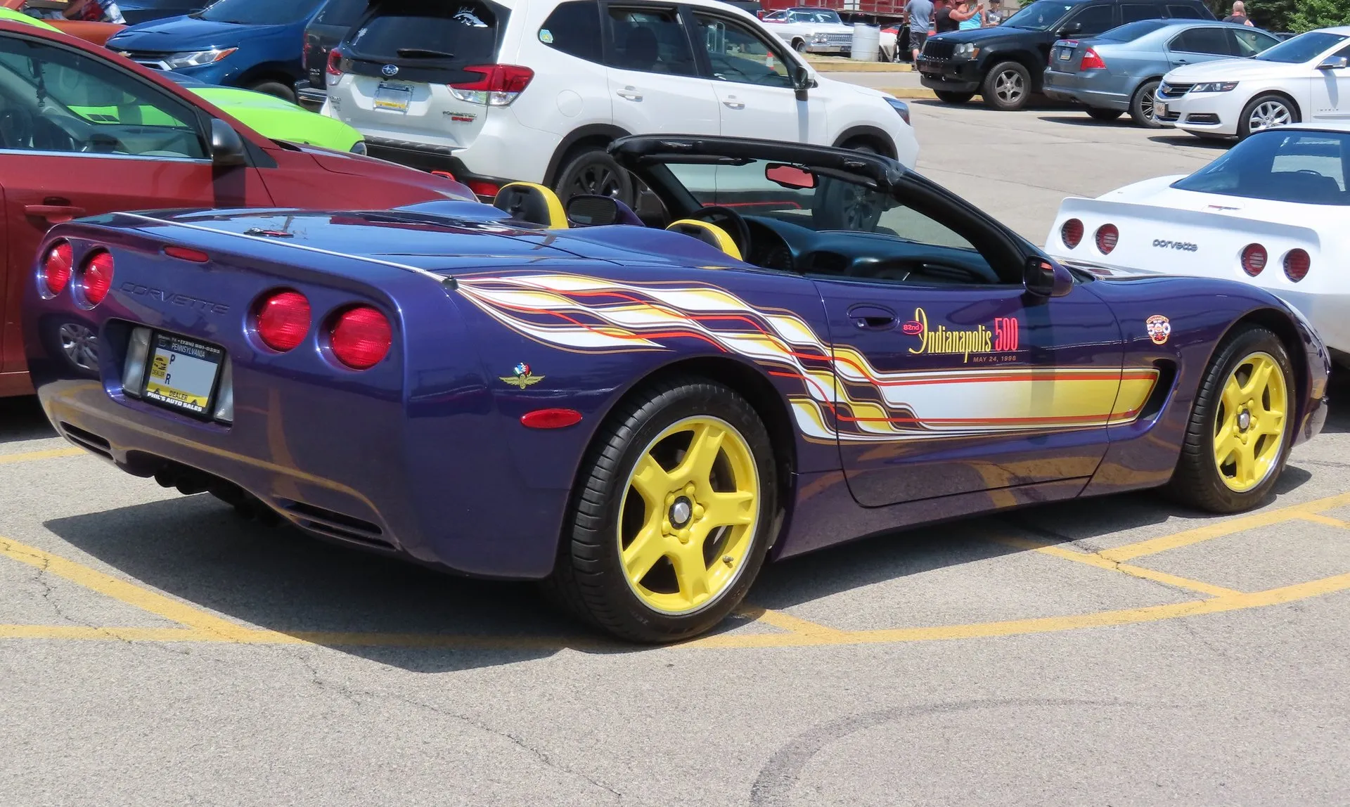 Purple C5 Corvette convertible with its top down in a parking lot, seen from the right rear, with checkered-flag style graphics along the side, Indianapolis 500 lettering dated May 24, 1998 on the door, yellow wheels and a Pennsylvania dealer plate.