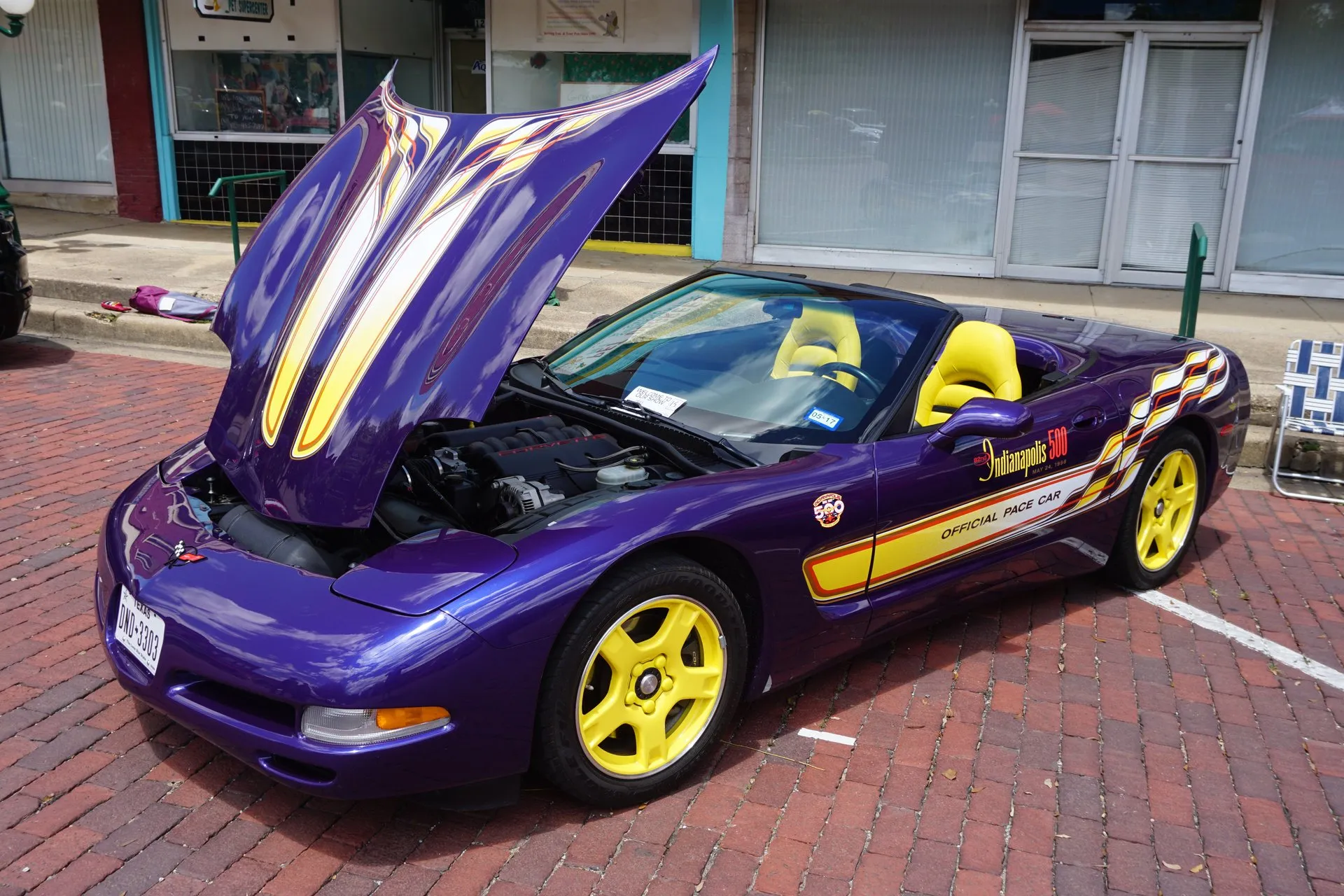 Purple C5 Corvette convertible with yellow and white graphics, yellow five-spoke wheels and yellow seats, parked on a brick street with its hood raised over the engine, an Indianapolis 500 Official Pace Car decal on the door and a Texas license plate.
