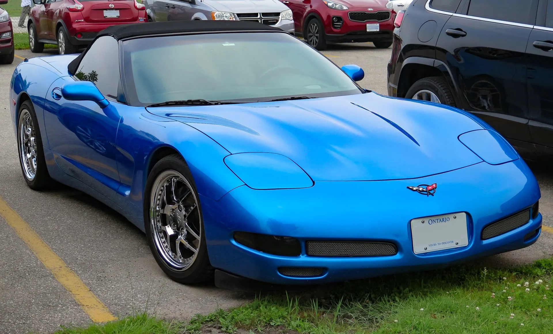 Bright blue C5 Corvette convertible with its black top up, parked at the edge of a lot beside grass, seen from the front right, on chrome multi-spoke wheels, with an Ontario license plate.