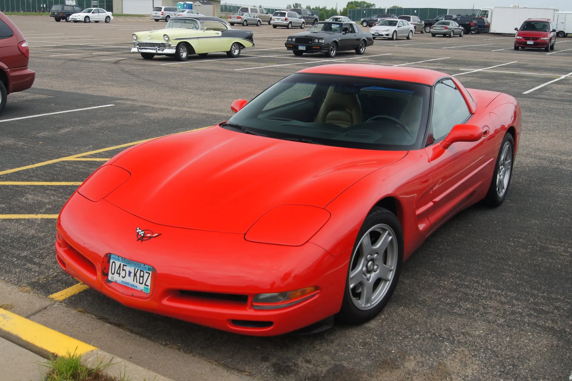 Red C5 Corvette coupe parked in a large lot, seen from the front left and above, with silver five-spoke wheels, closed pop-up headlights, a tan interior and a Minnesota license plate, and a two-tone 1950s Chevrolet behind it.