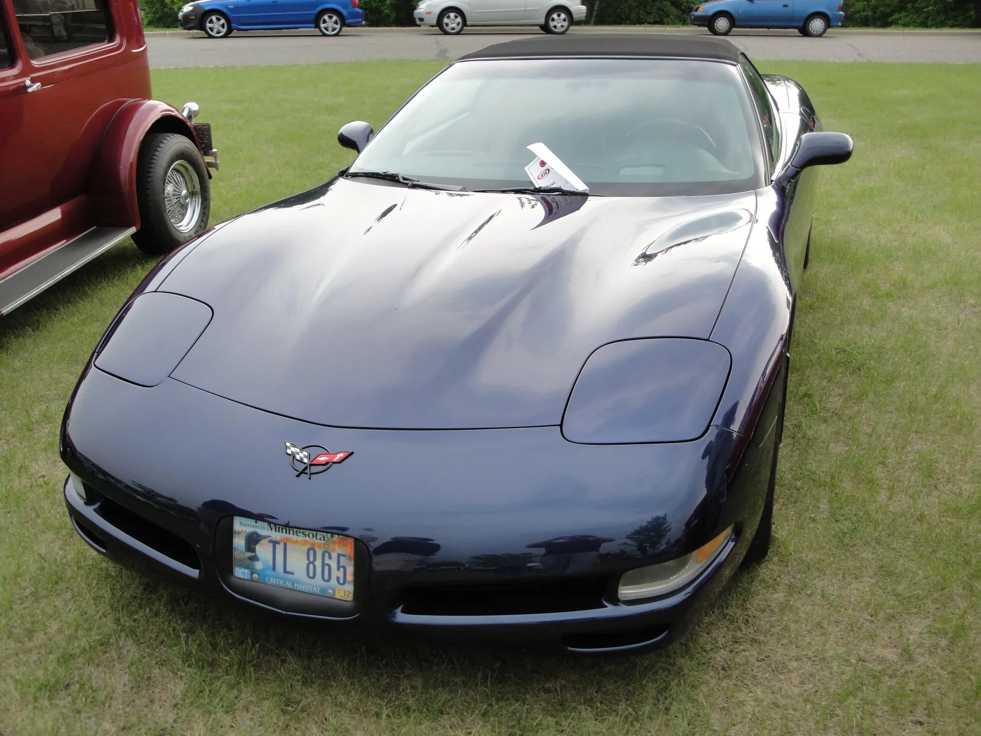 Dark blue C5 Corvette convertible with its black top up, parked on grass at a car show and seen from the front, with a show card on the dashboard and a Minnesota license plate.