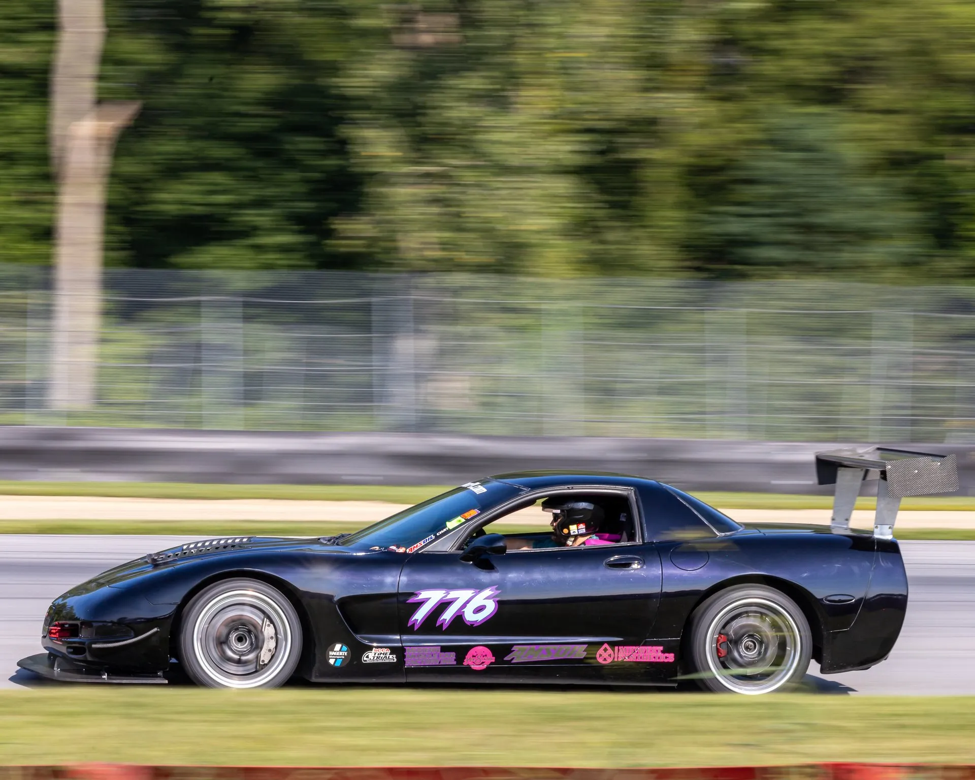 Black, heavily modified C5 Corvette with a tall rear wing, a front splitter and a vented hood, at speed on a race track, seen from the left side, with the number 776 on the door and a helmeted driver.