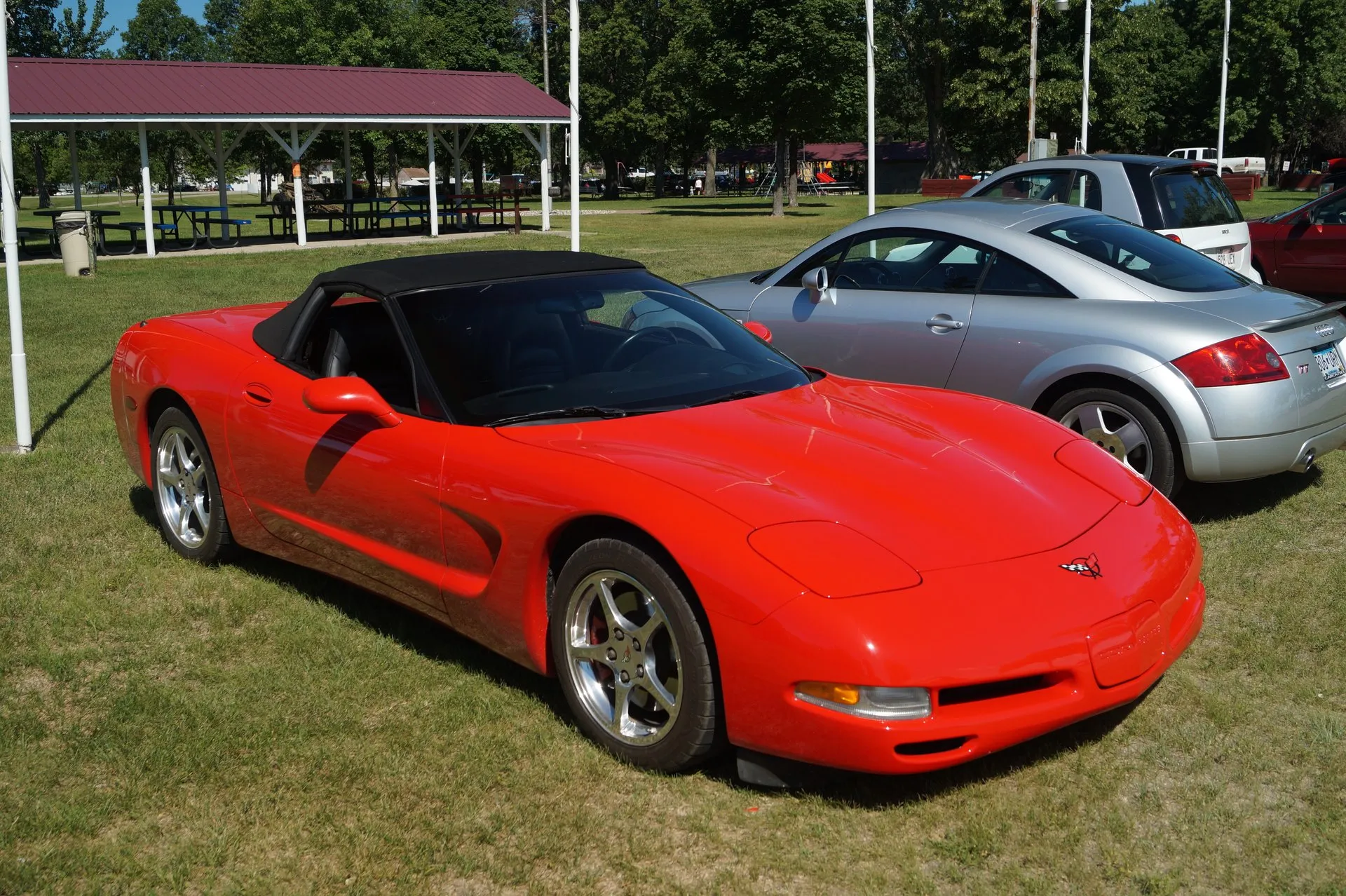 Red C5 Corvette convertible with its black top up, parked on grass at a car show and seen from the front right, with silver five-spoke wheels and a silver Audi TT parked behind it.