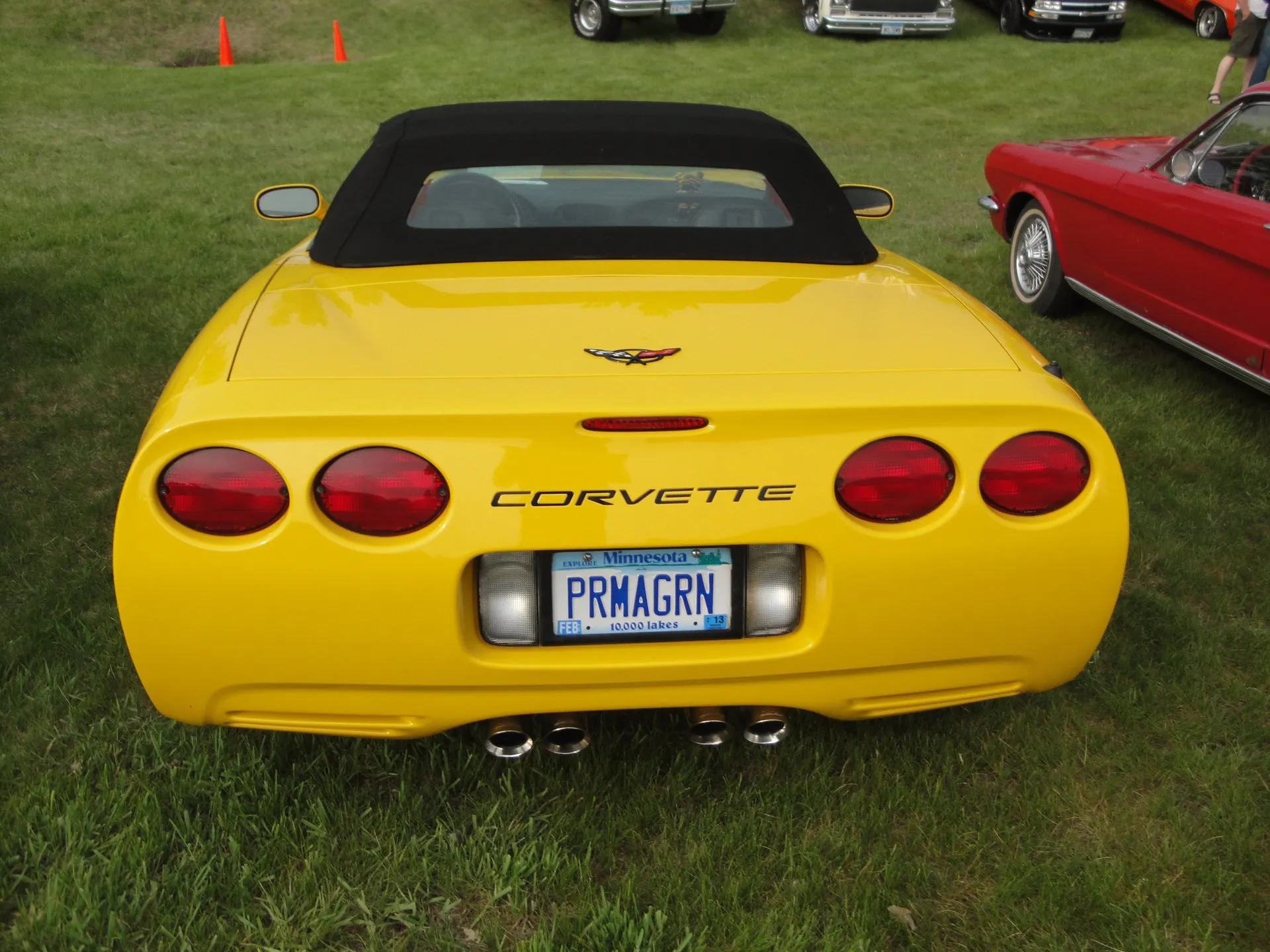 Yellow C5 Corvette convertible with its black top up, seen squarely from behind on grass, showing four round taillights, Corvette lettering, a Minnesota plate and four chrome exhaust tips.