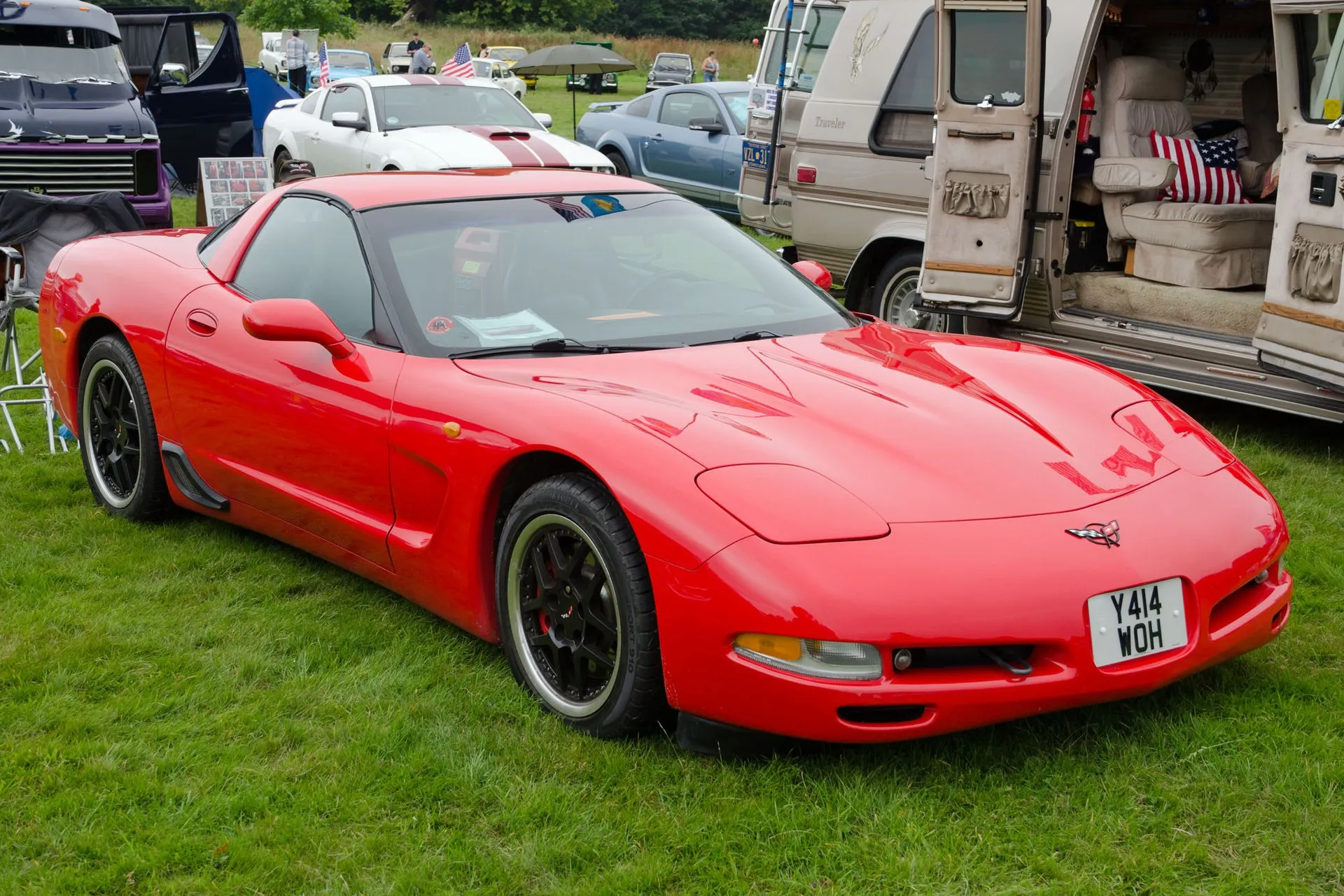 Red C5 Corvette coupe parked on grass at a car show, seen from the front right, with black split-spoke wheels and a British registration plate, a camper van with its doors open beside it.
