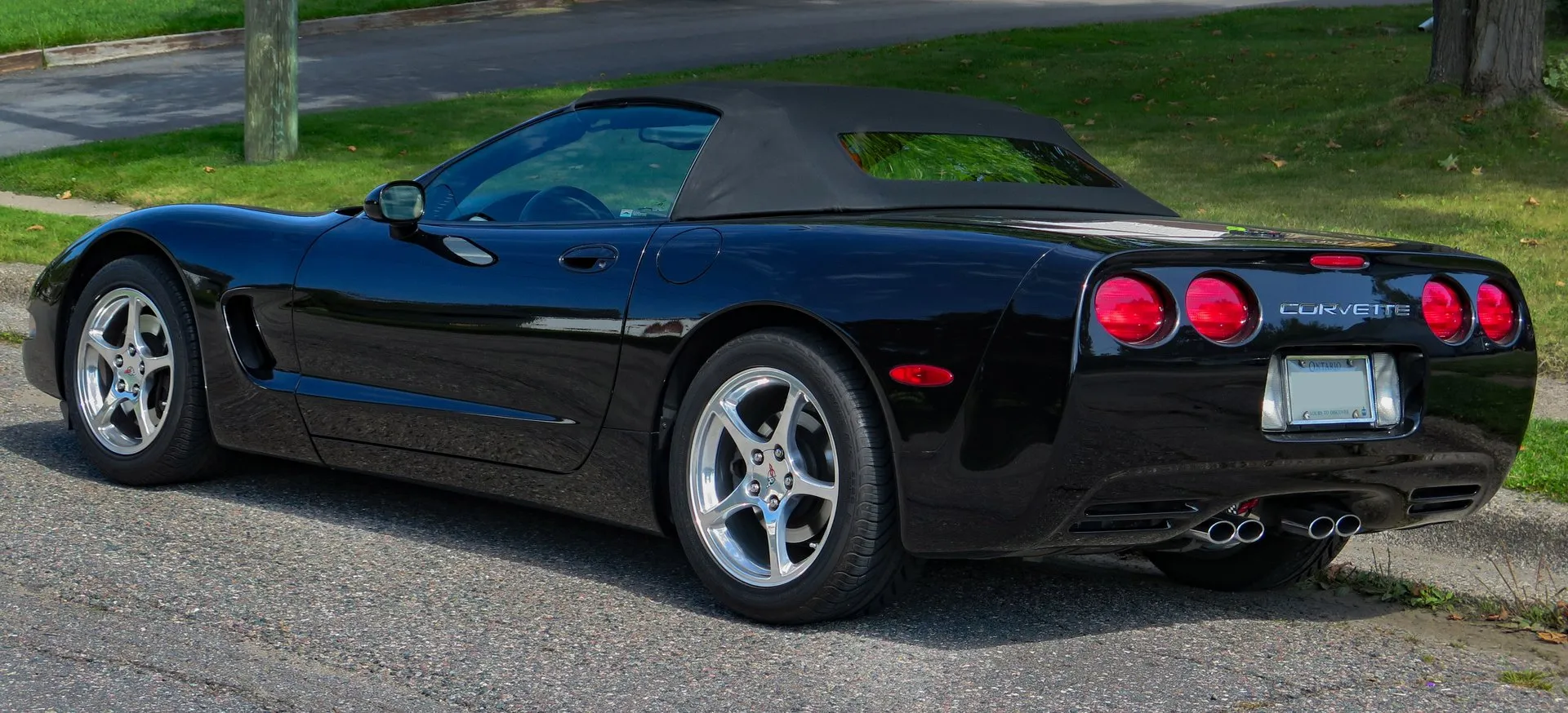 Black C5 Corvette convertible with its black soft top up, parked at a curb and seen from the rear left, showing four round taillights, four exhaust tips and polished five-spoke wheels.
