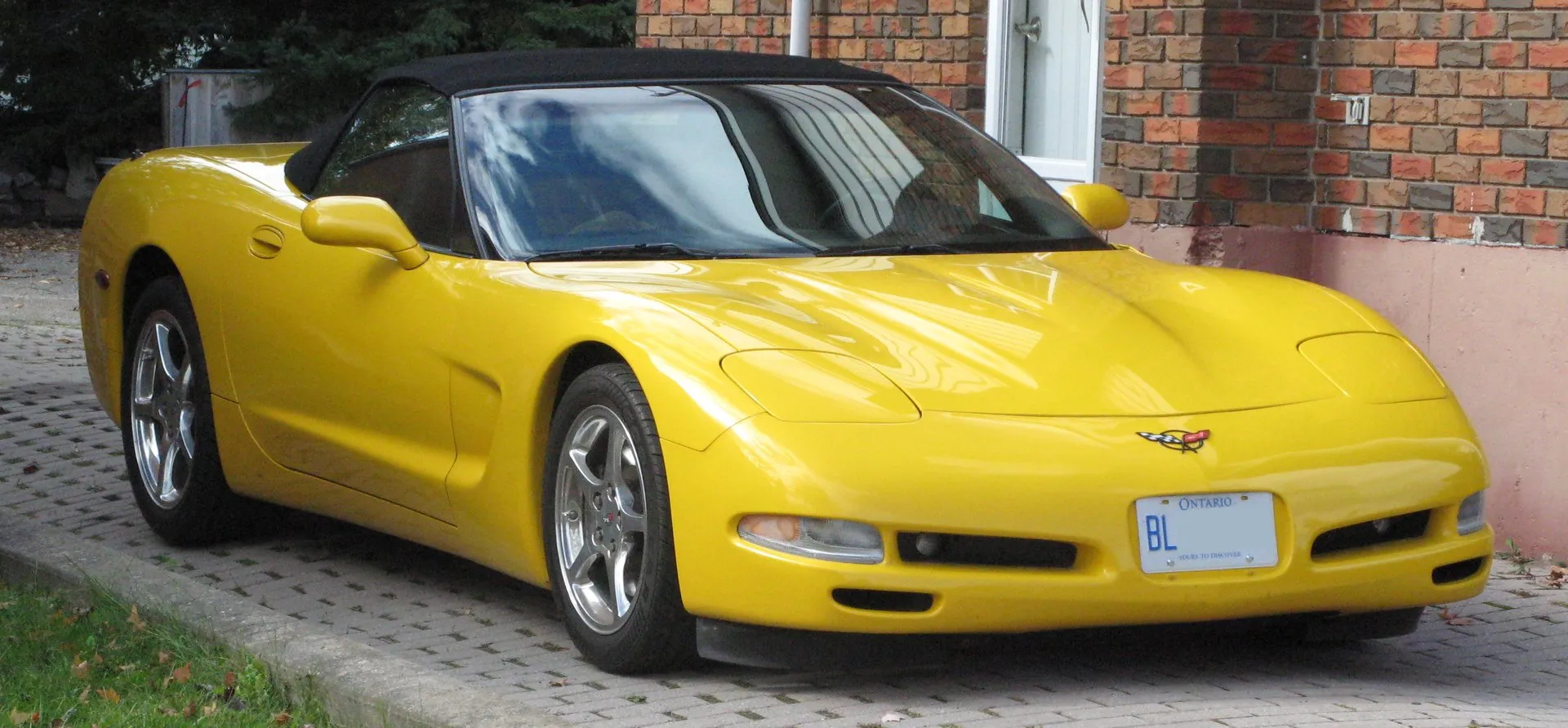 Yellow C5 Corvette convertible with its black top up, parked on a paved driveway in front of a brick house, seen from the front right, with silver five-spoke wheels.