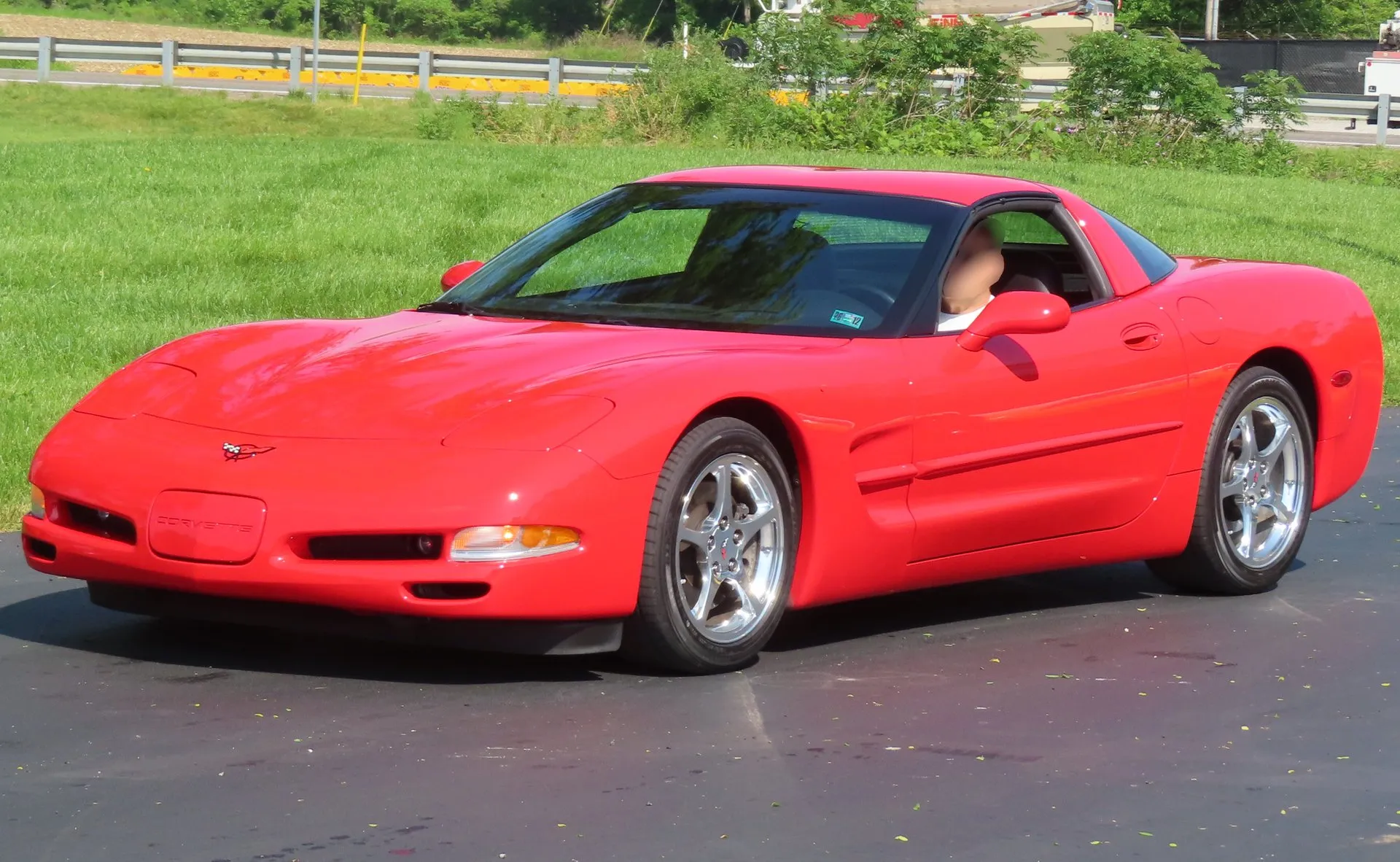 Torch Red C5 Corvette coupe on an asphalt lot beside grass, seen from the front left, with polished five-spoke wheels, a crossed-flags emblem on the nose and a driver in the seat.