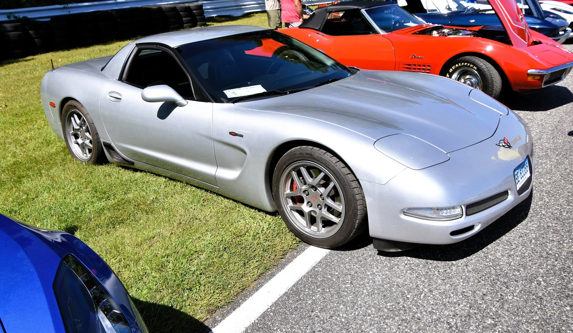 Silver C5 Corvette Z06 parked on grass at a car show, seen from the front right, with gray five-spoke Z06 wheels, red brake calipers and a fixed roof, a red C3 Corvette with its hood open behind it.