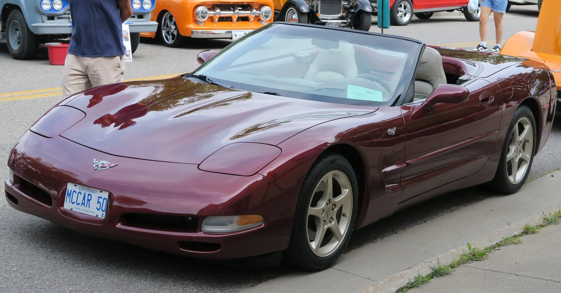 Dark red 2003 Corvette 50th Anniversary Edition convertible with the top down, parked at a street car show and seen from the front left, with a light tan interior, champagne five-spoke wheels and a 50 badge on the front fender.