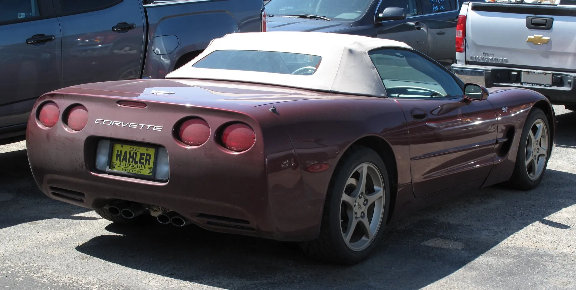 Dark red 2003 Corvette 50th Anniversary Edition convertible with a tan soft top raised, parked in a lot and seen from the rear right, with four round taillights and a dealer plate.