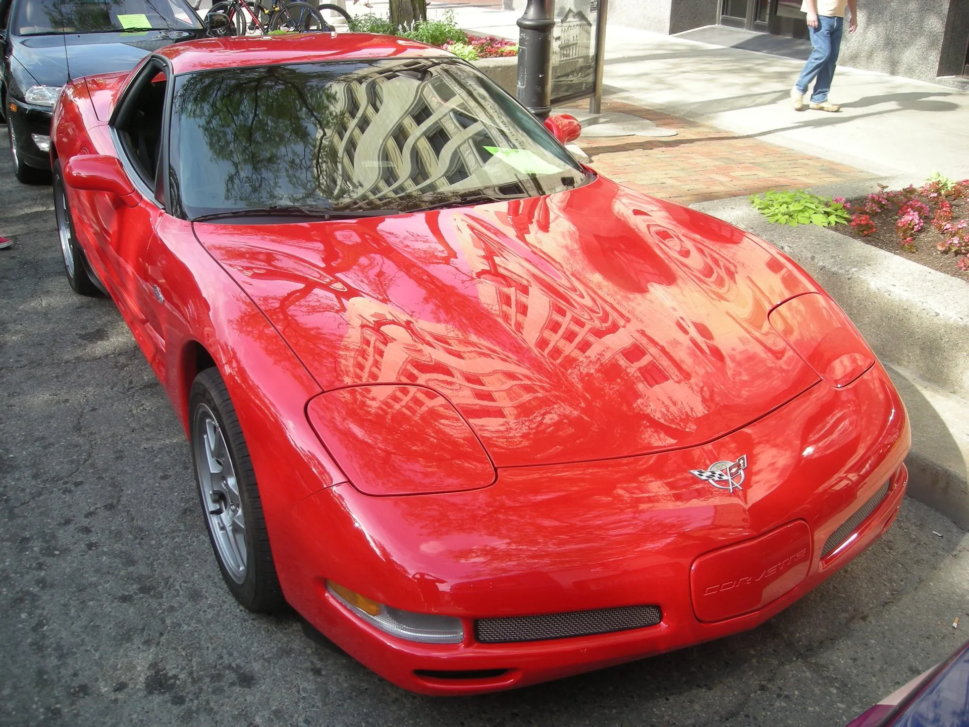 Red 2003 Chevrolet Corvette Z06 coupe parked on a city street beside a planter, seen from above the front right, with mesh grille inserts and silver wheels.