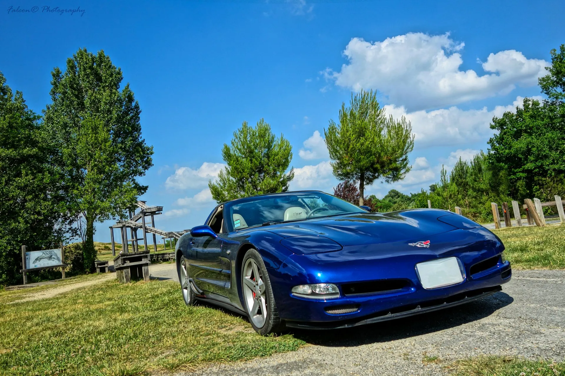 Le Mans Blue C5 Corvette coupe on a gravel path beside grass and trees, seen low from the front right, with light-colored seats and an embroidered headrest behind the windshield, silver five-spoke wheels and a blank front plate.