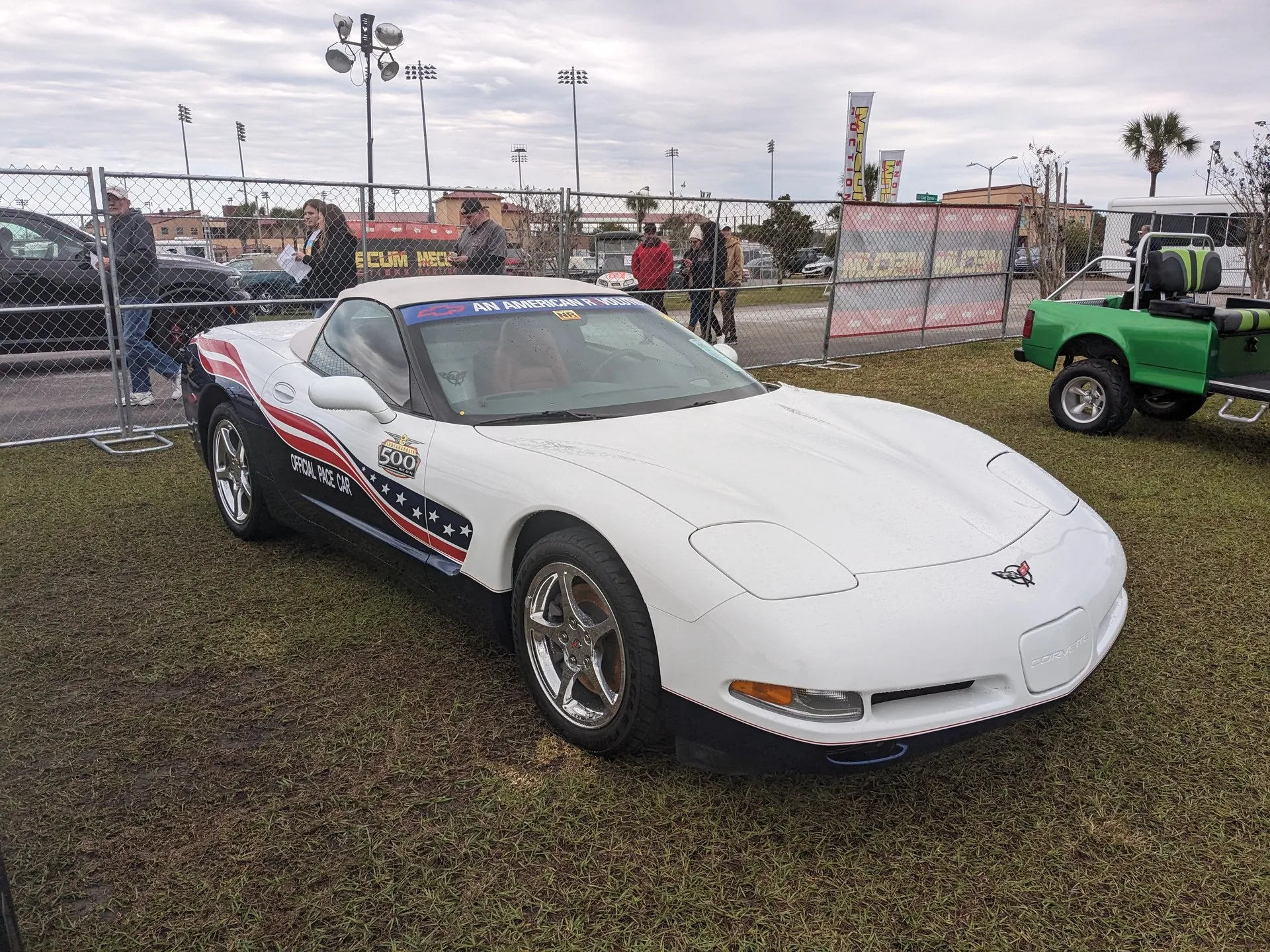White C5 Corvette convertible with its light top up, parked on grass in front of a chain-link fence at an auction, seen from the front right, with dark blue lower sides, red, white and blue stripes and stars, an Official Pace Car decal, an Indianapolis 500 logo and An American Revolution across the top of the windshield.