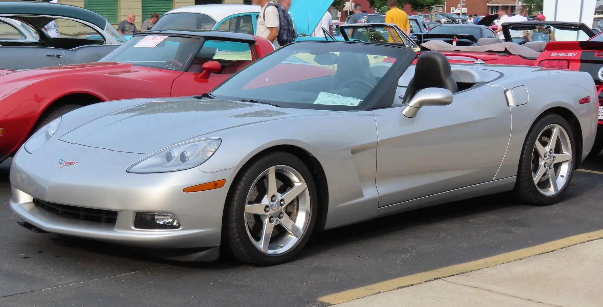 Silver Chevrolet Corvette C6 convertible with the top down, parked at an outdoor car show and seen from the front left, with polished five-spoke wheels.