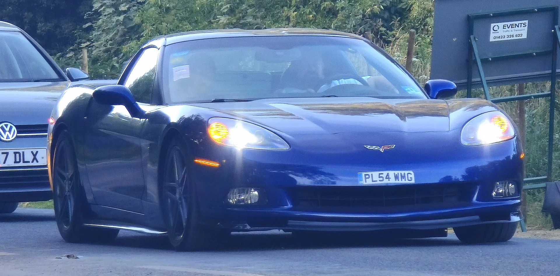 Dark blue Chevrolet Corvette C6 coupe with its headlights on, seen from the front on a road lined with bushes, with dark wheels and a British registration plate.
