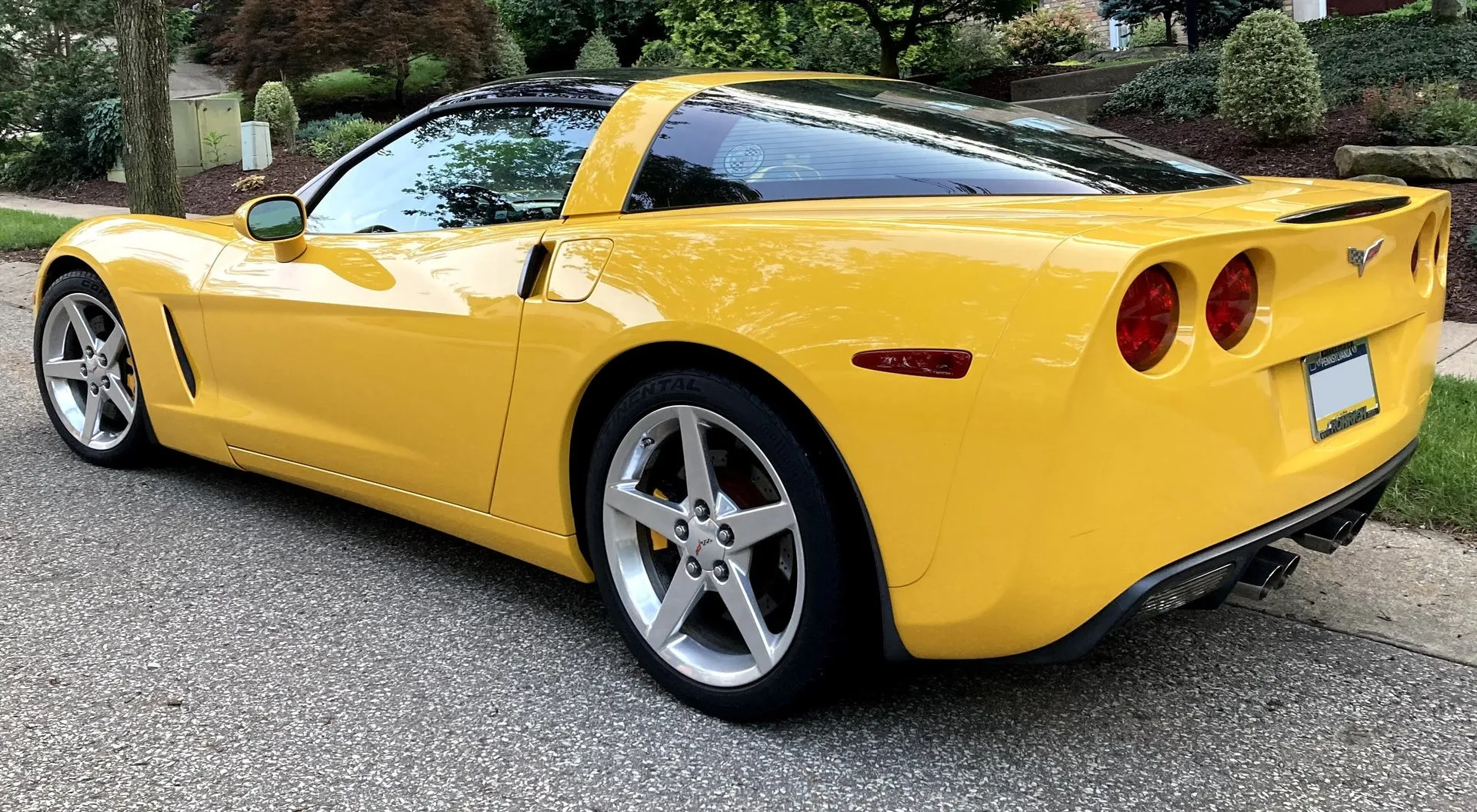 Yellow Chevrolet Corvette C6 coupe with a black roof panel, seen from the rear left on a residential street, showing four round taillamps and four exhaust tips in the center.