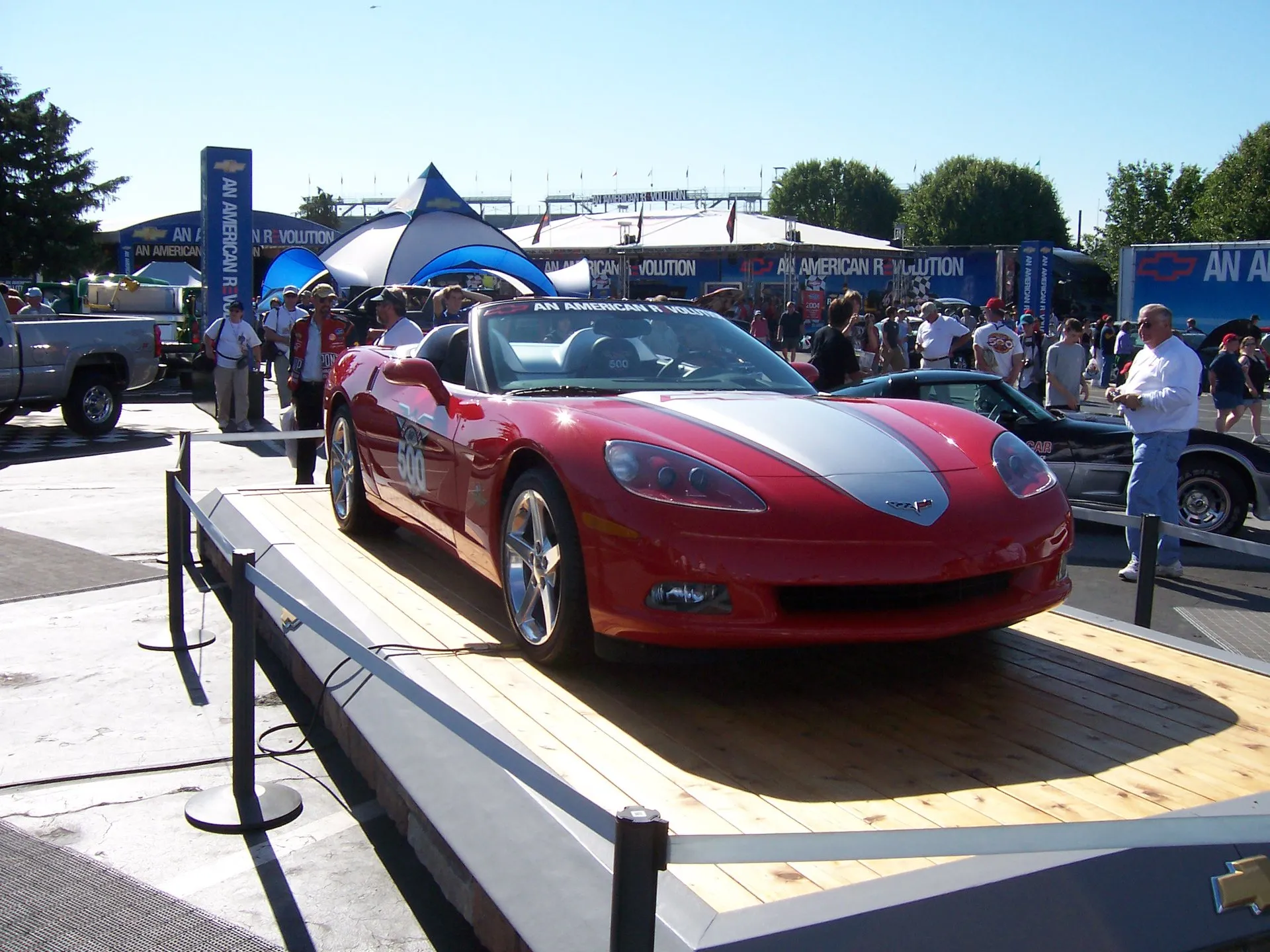 Red Chevrolet Corvette C6 convertible with a wide silver center stripe and 500 logos on the doors, roped off on a wooden display stage, with crowds and Chevrolet banners behind.