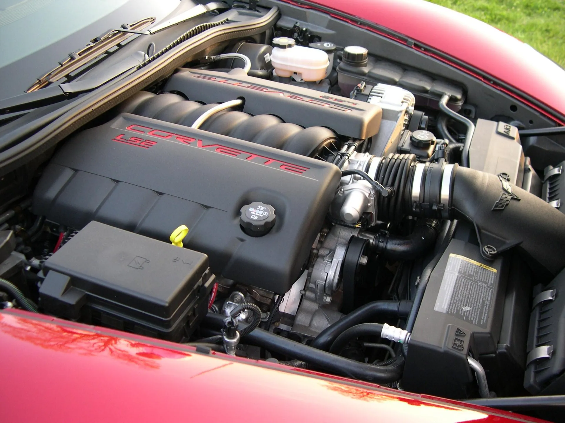 Open engine bay of a red C6 Corvette, showing a black engine cover lettered Corvette LS2 in red, the air intake with a crossed-flags emblem and a black fuse box in the foreground.