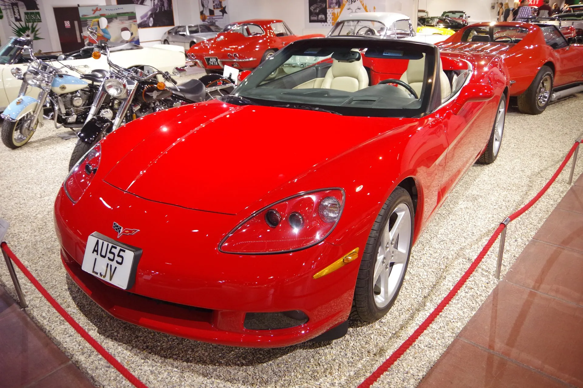 Red C6 Corvette convertible with the top down and light tan seats, displayed on gravel behind a red rope in a museum hall, seen from the front left, with a British registration plate.