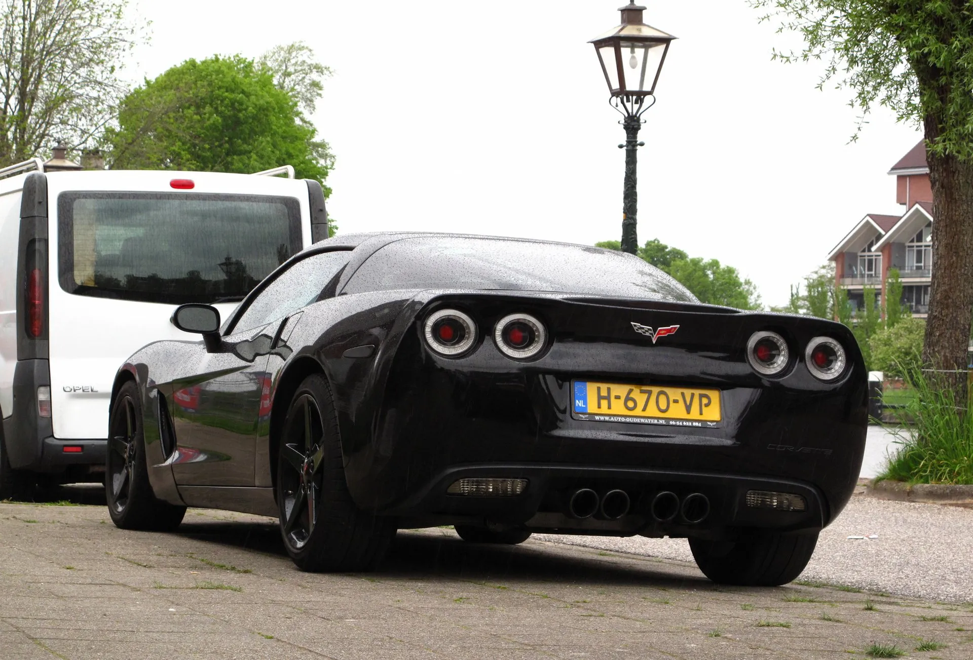 Black C6 Corvette coupe with black wheels on a wet brick-paved street beside a white van, seen from the rear left, showing four round taillamps, four exhaust tips in the center and a Dutch plate.