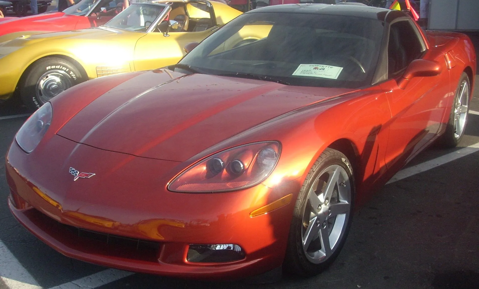 Orange metallic C6 Corvette coupe with a dark roof panel and silver five-spoke wheels at an outdoor car show, seen from the front left, with a show card on the dashboard.