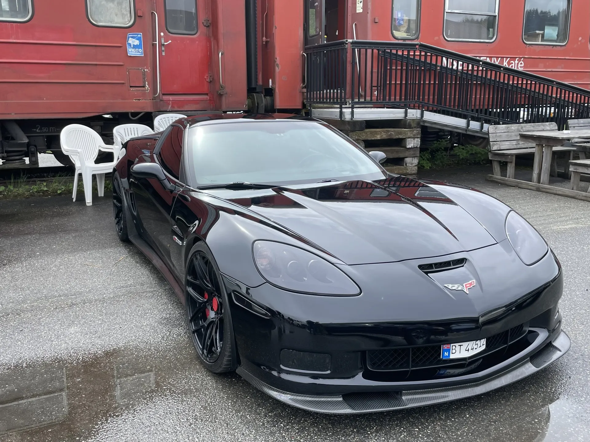 Black C6 Corvette Z06 coupe with black wheels, red brake calipers and a carbon-fiber front splitter, parked on wet pavement beside old red railway carriages, with a Norwegian plate.