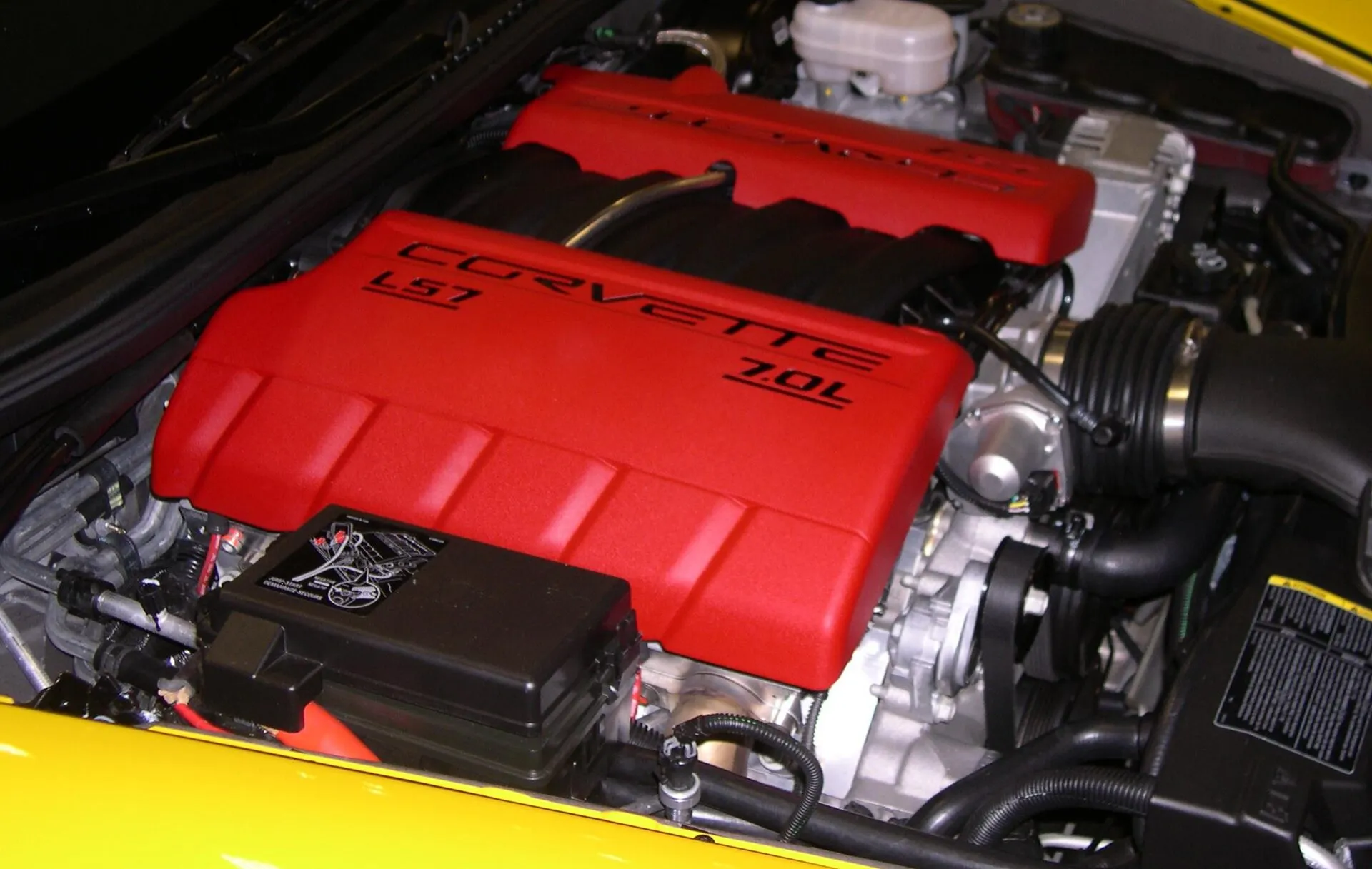 Engine bay of a yellow C6 Corvette Z06 with a red engine cover lettered Corvette, LS7 and 7.0L; in the foreground, the black underhood fuse block with a printed jump-start diagram.