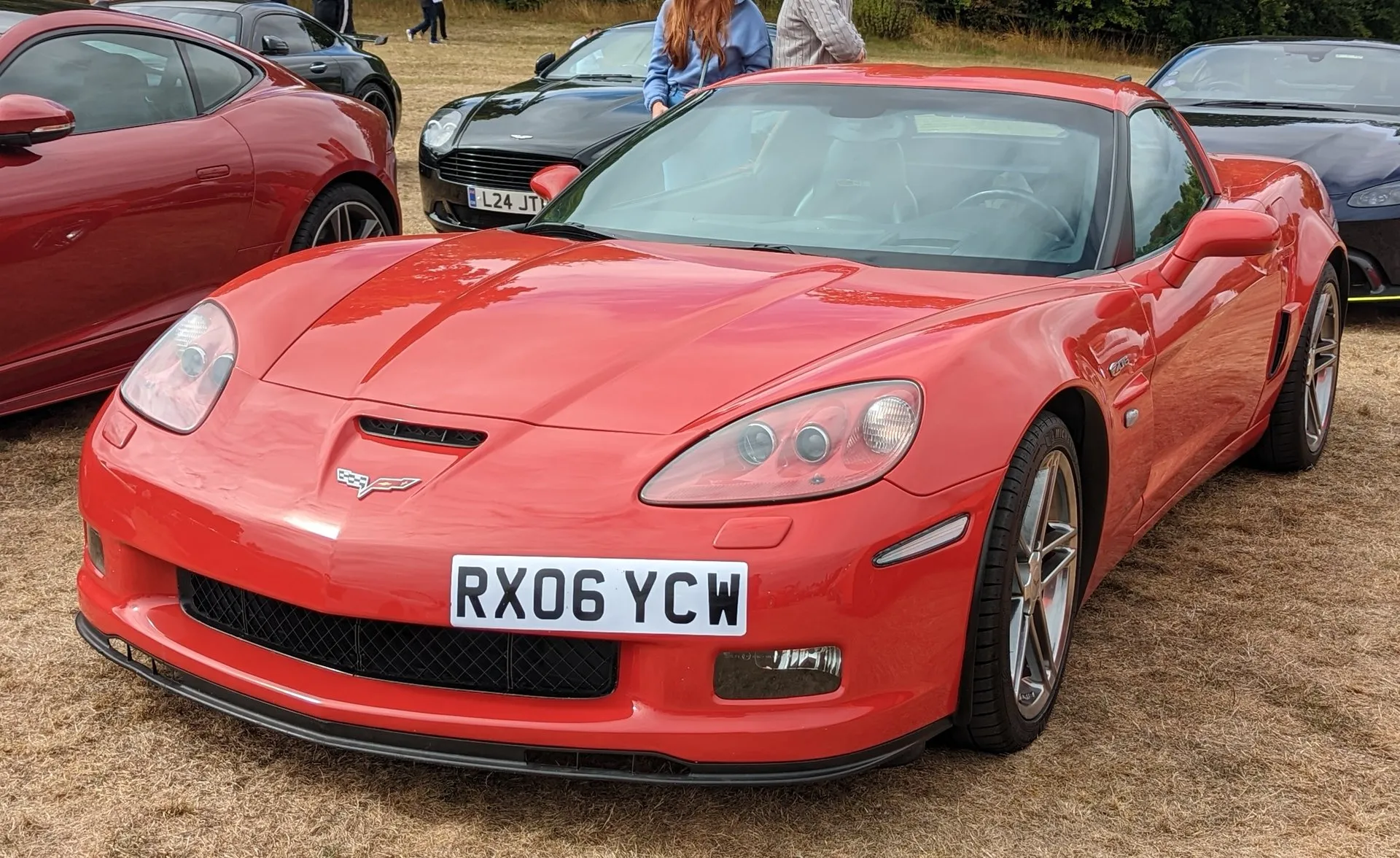 Red C6 Corvette Z06 coupe parked on dry grass at a car show, seen from the front left, with a hood vent, a front splitter, a Z06 badge on the front fender and a British plate.