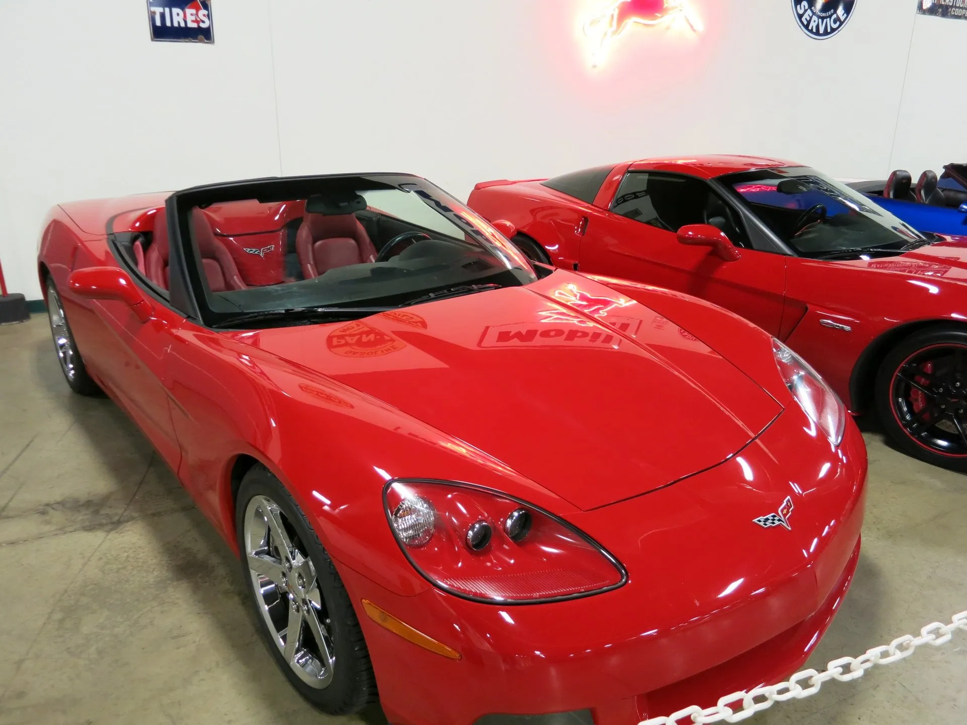 Red C6 Corvette convertible with the top down, red leather seats and chrome five-spoke wheels, parked indoors in a museum beside another red C6, with old service-station signs on the white wall.