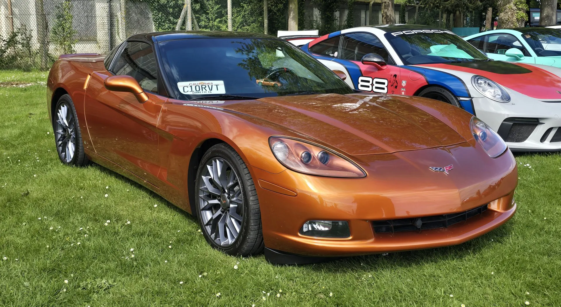 Orange metallic C6 Corvette coupe with a dark roof panel and grey multi-spoke wheels on grass at a car show, seen from the front right, with a California plate in the windshield and Porsches parked beside it.