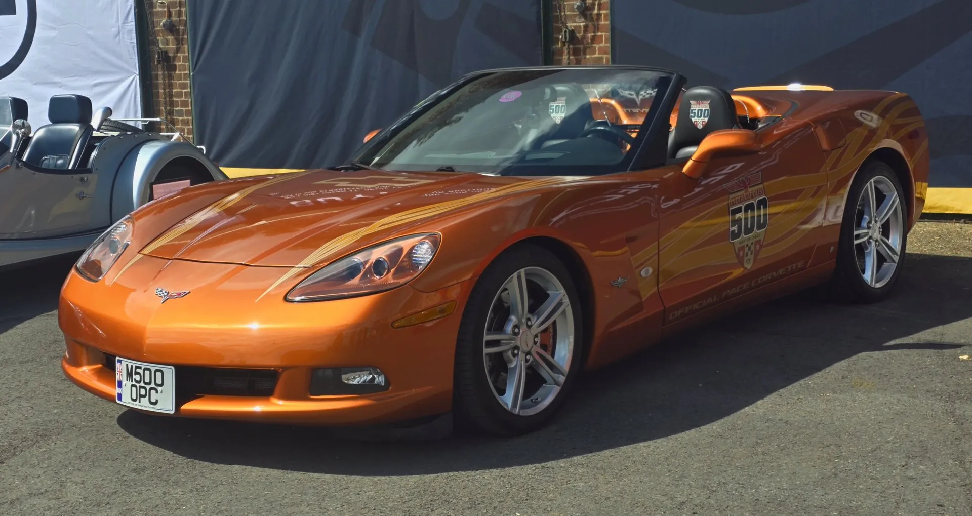 Orange C6 Corvette convertible with the top down, gold ribbon graphics across the hood and door, an Indianapolis 500 logo and Official Pace Corvette lettering on the door, and silver split-spoke wheels, parked on tarmac at a British show.