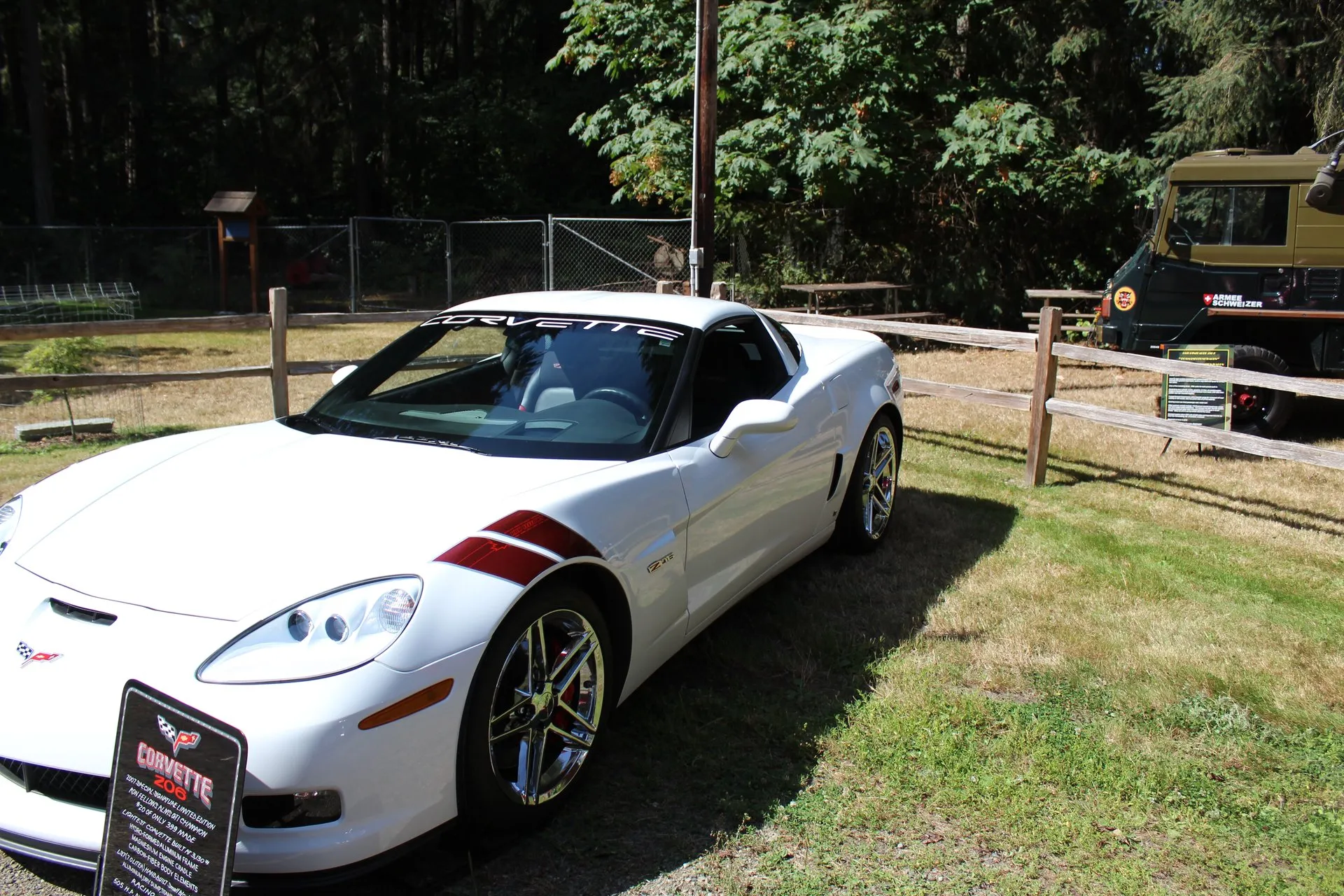 White C6 Corvette Z06 coupe with two dark red stripes across the front fender, chrome wheels and a Corvette windshield banner, parked on grass by a wooden fence, with a display placard in front of it.