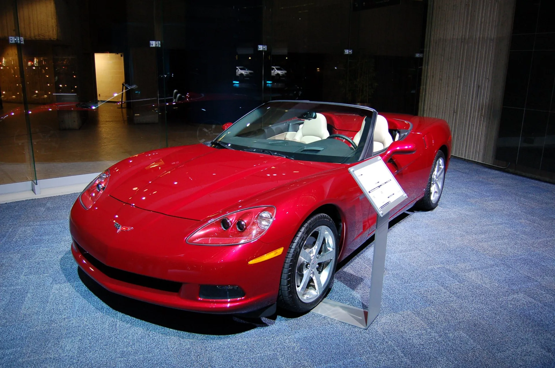 A red Corvette C6 convertible with its top down and a light interior, displayed on a blue carpeted platform in a showroom with a placard stand beside it.