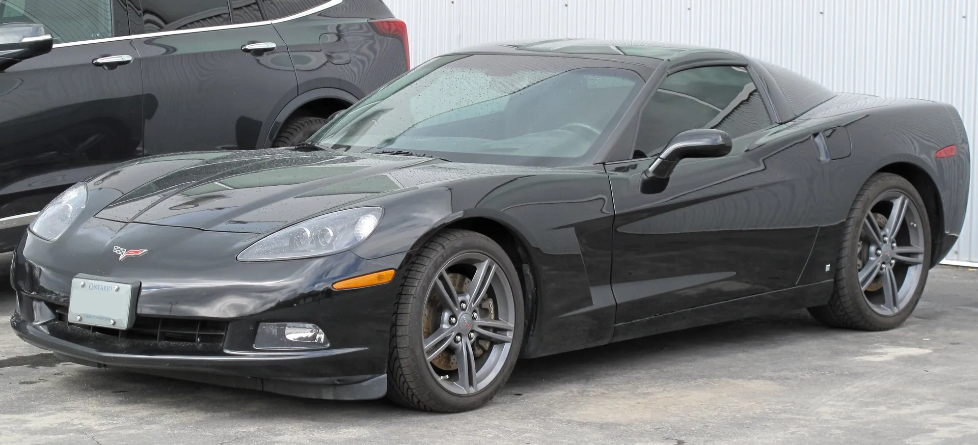 A black Corvette C6 coupe with dark split-spoke wheels, seen from the front left corner in a parking lot.